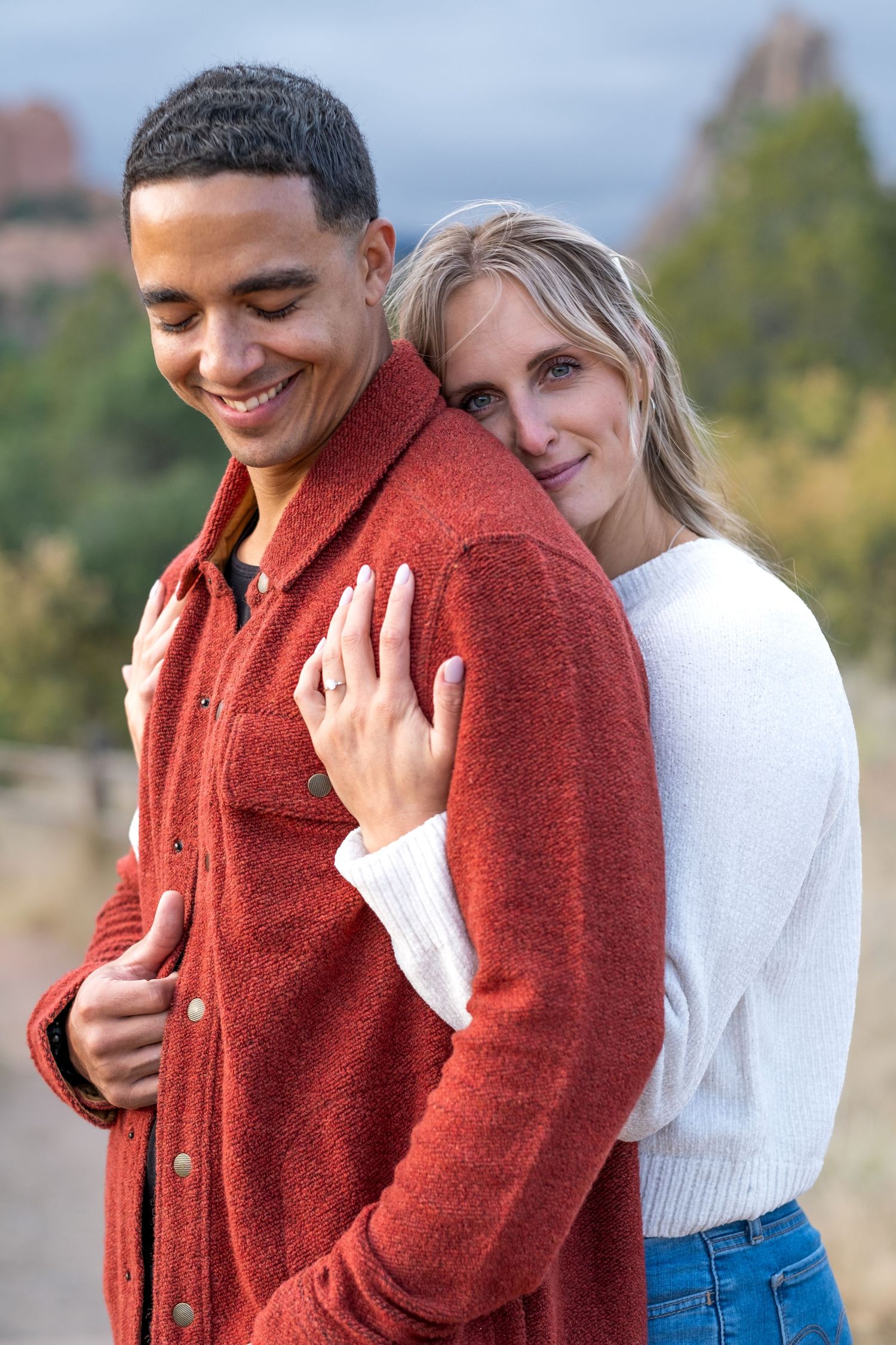 A smiling interracial couple embraces outdoors, with the woman resting her head on the mans shoulder in nature.