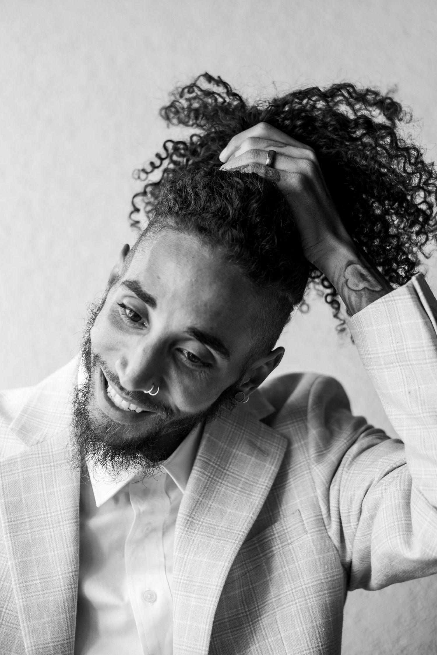 Stylish groom in plaid blazer running hand through curly hair, smiling in black and white portrait.