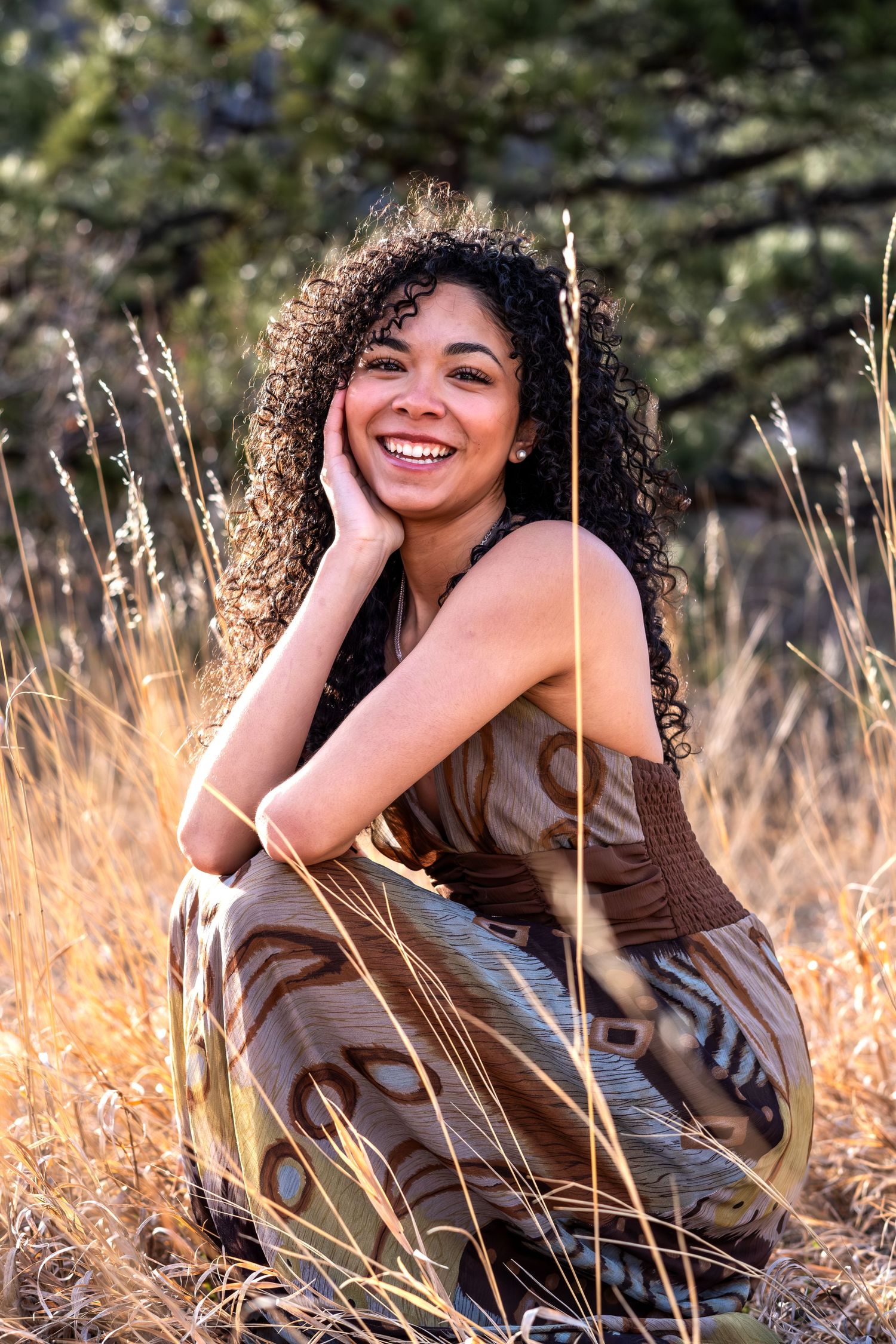 Happy woman with curly hair in bohemian dress sitting in tall golden grass outdoors.