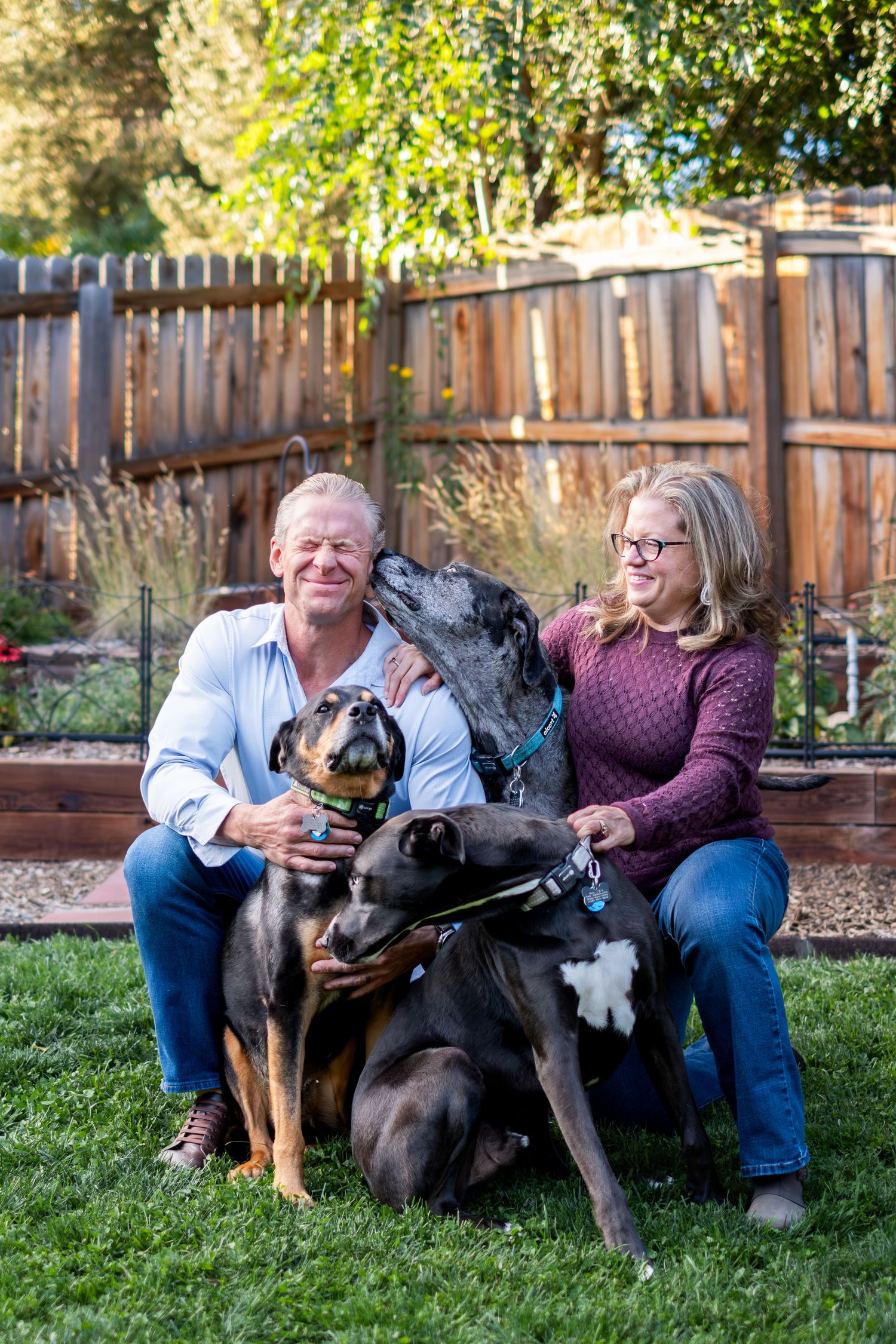 Smiling couple sits on grass with two playful black dogs in a fenced backyard garden.