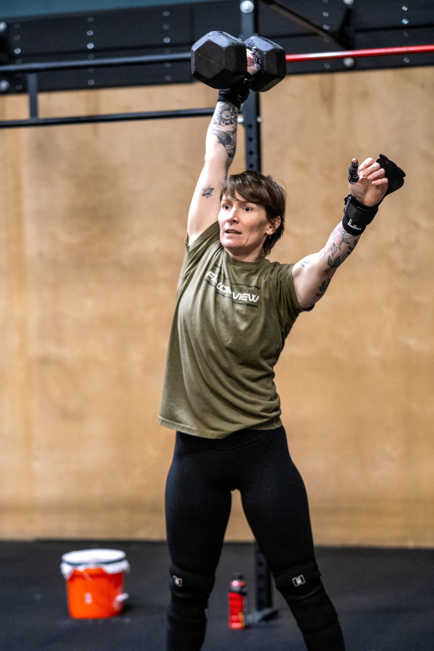 Tattooed woman in olive shirt performs single-arm overhead dumbbell press in CrossFit gym.
