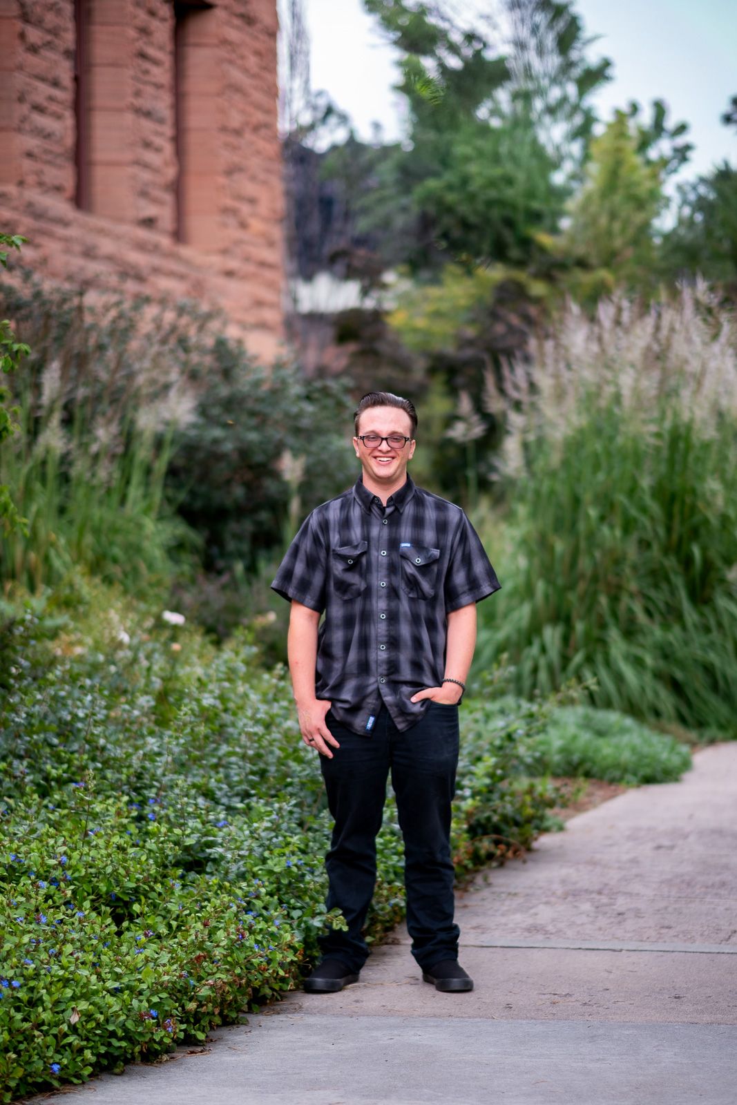 Smiling young man in plaid shirt standing on a garden path beside lush greenery and brick building.