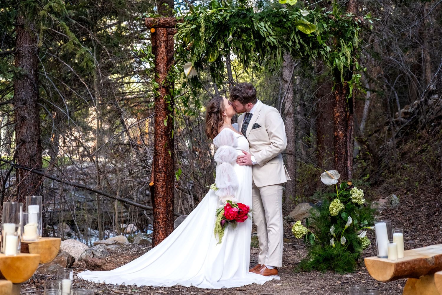 Newlyweds sharing first kiss under a lush greenery-covered wooden arch in an outdoor forest wedding ceremony.