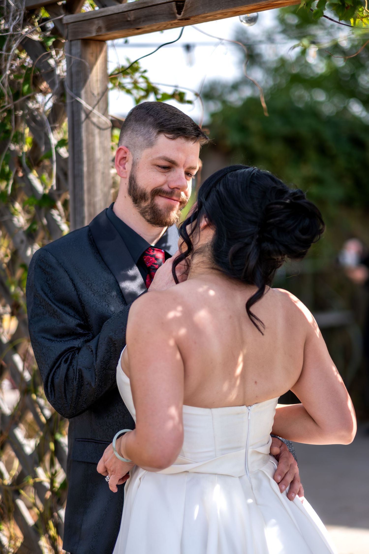Groom in black suit smiling at bride in white strapless wedding dress under a wooden arbor.