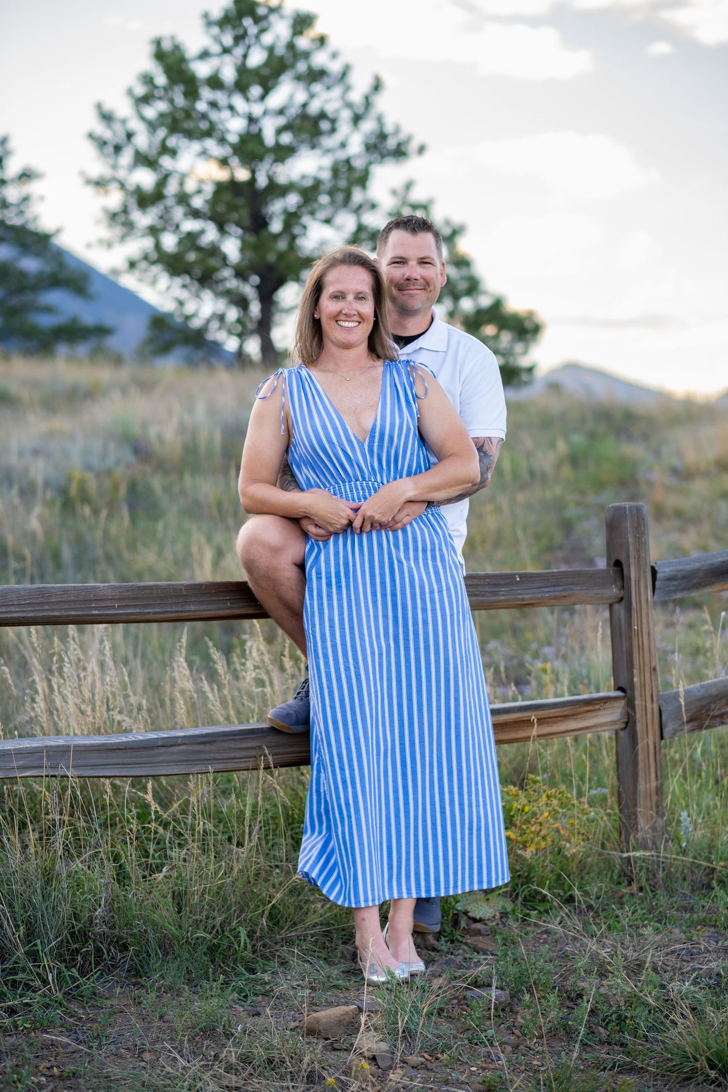 Smiling couple posing by a rustic wooden fence in a mountain meadow, woman in blue striped dress.