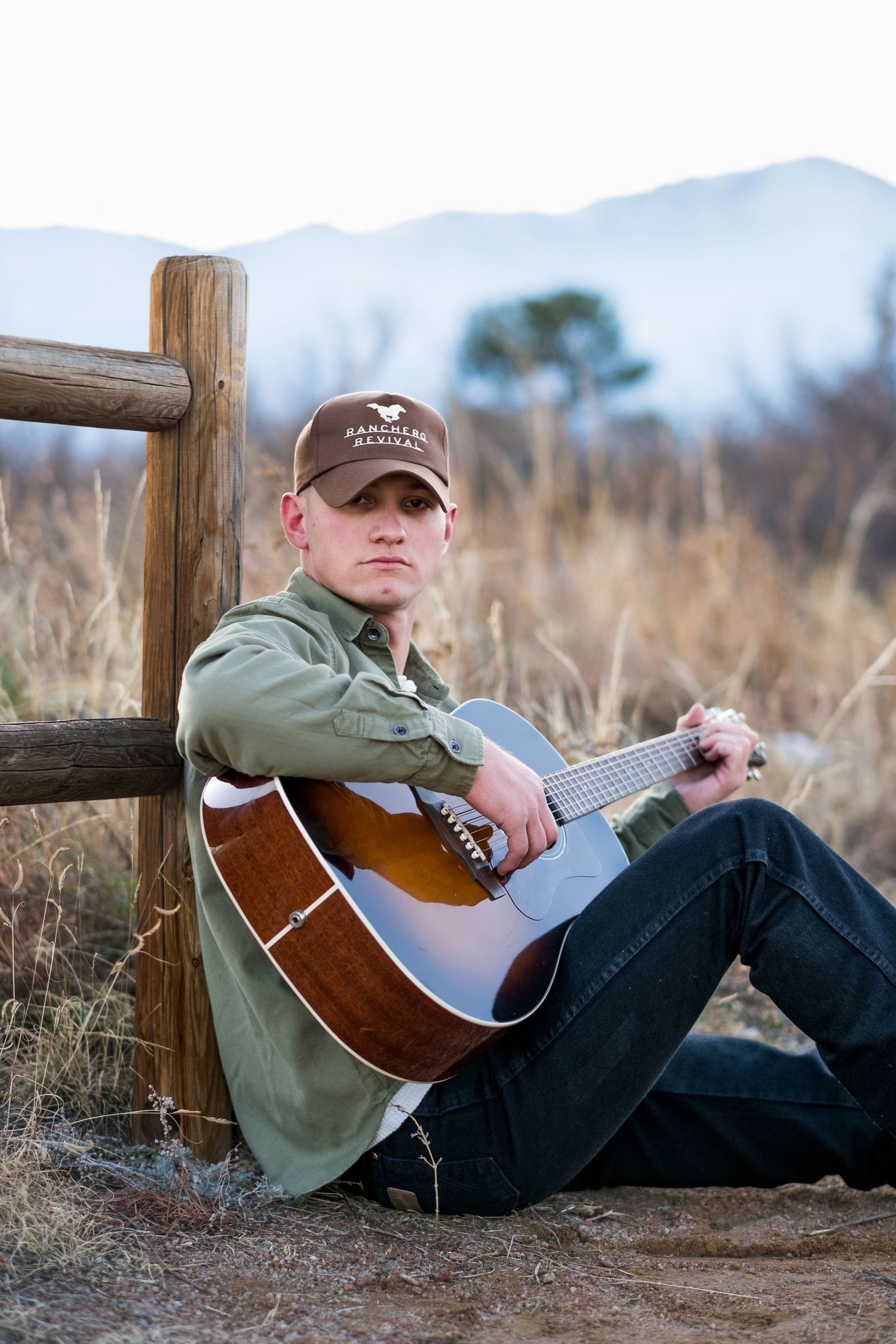 Young male guitarist in brown cap sitting against wooden fence playing acoustic guitar at dusk.