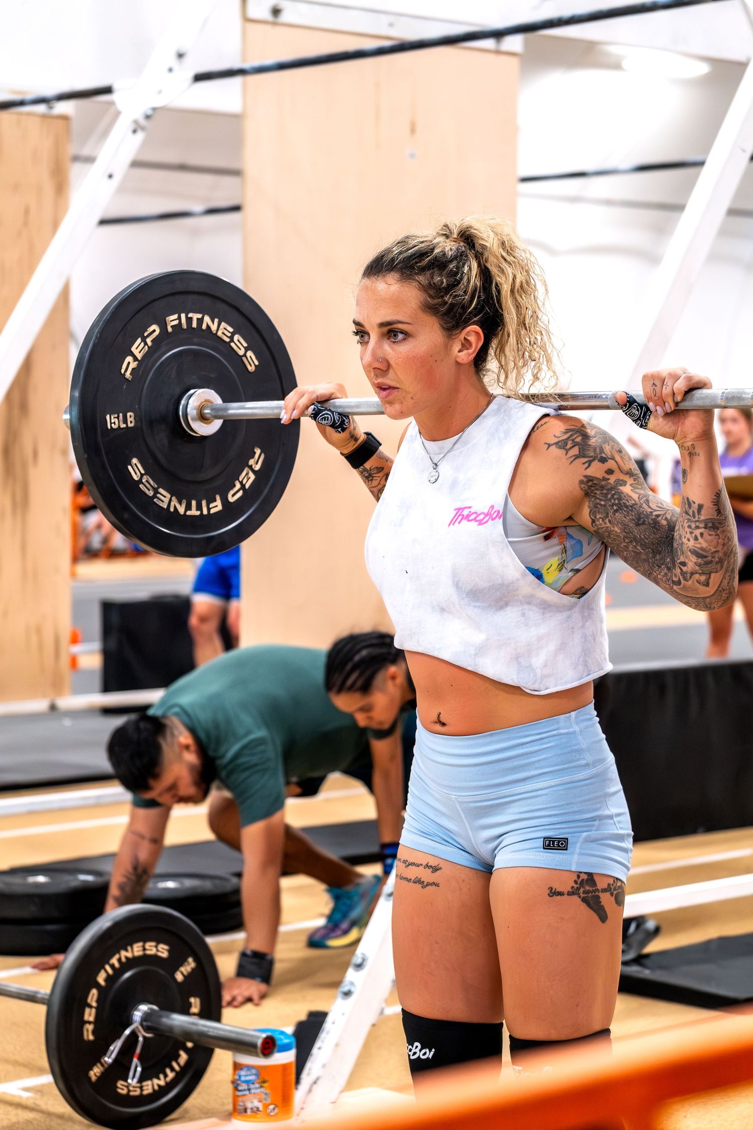 Tattooed woman in athletic wear preparing to back squat a loaded barbell at a CrossFit competition.