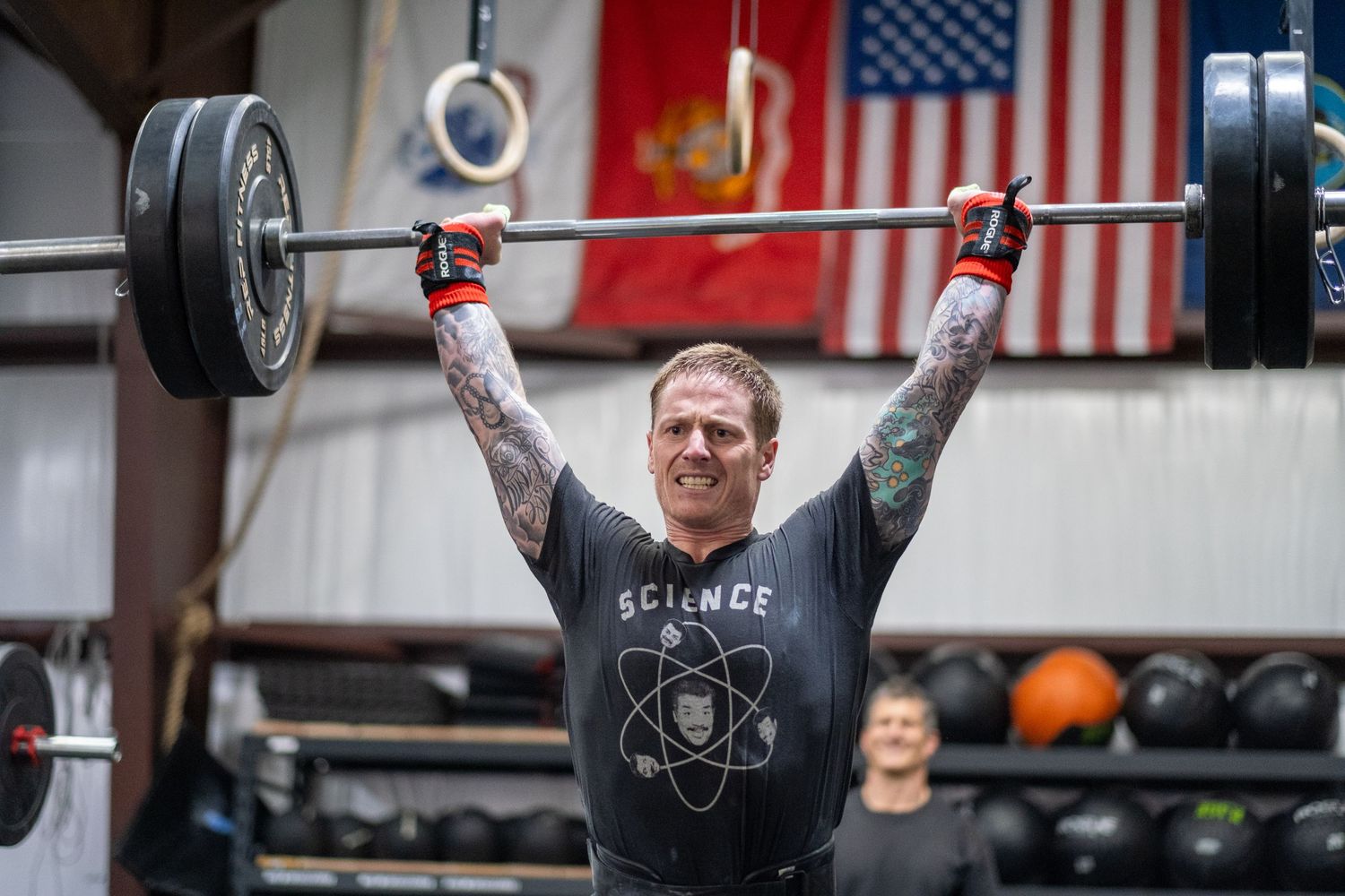 Tattooed man overhead pressing a loaded barbell in a gym with flags on the wall.