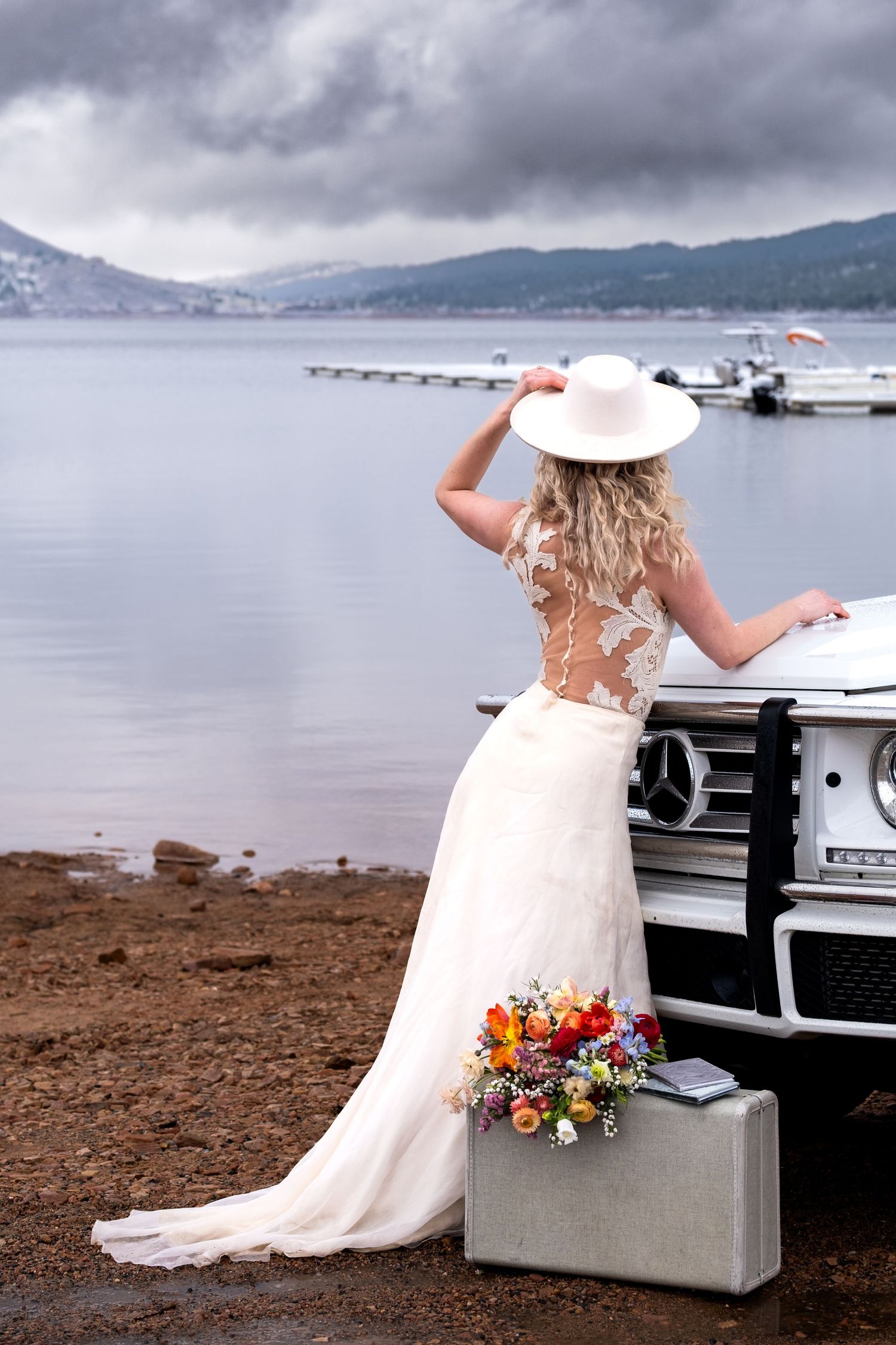 Bride in white lace gown and hat leans on Mercedes SUV by lake with colorful bouquet and vintage suitcase.