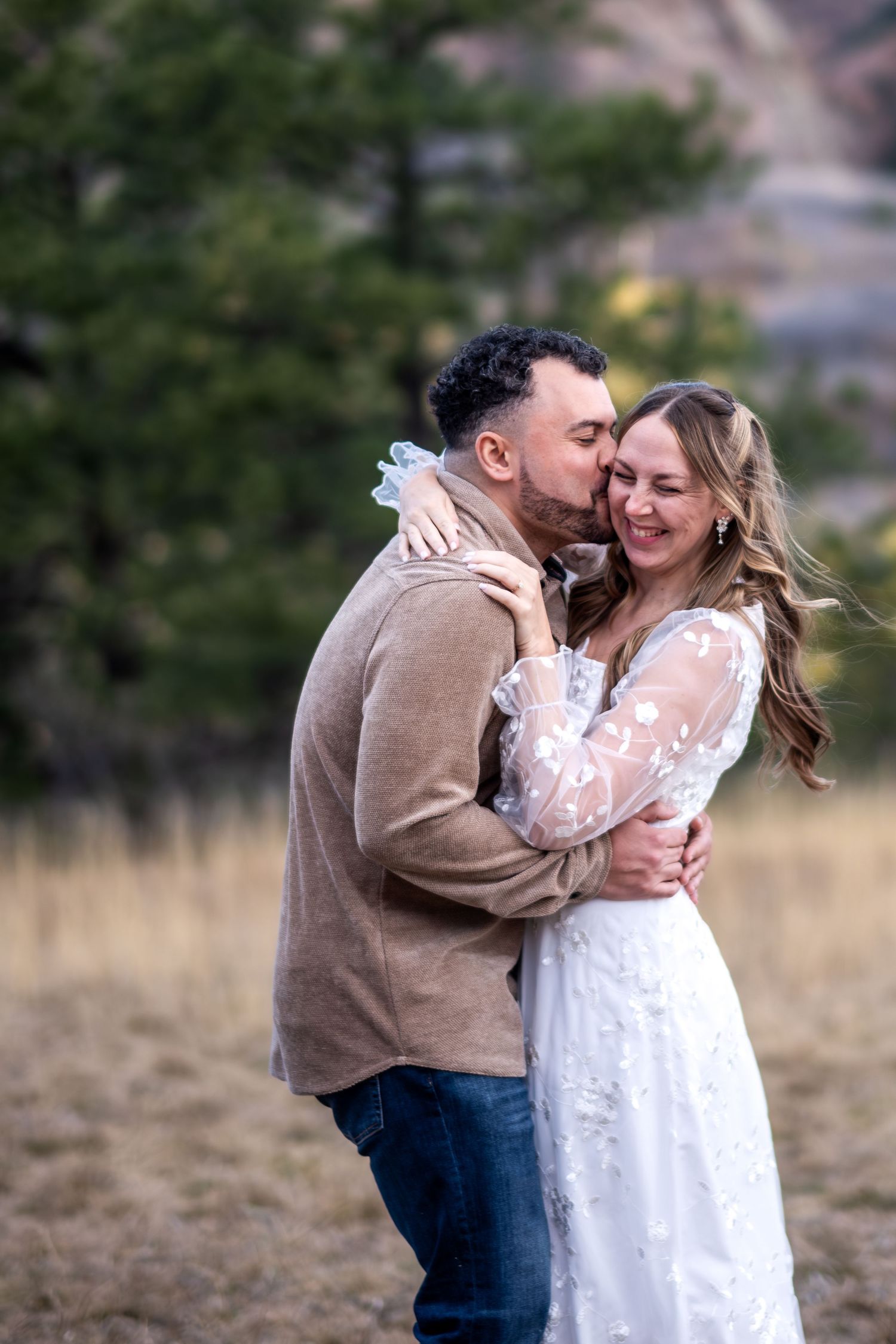 Couple embracing in a field, groom kissing bride's cheek as she smiles in a white lace wedding dress.
