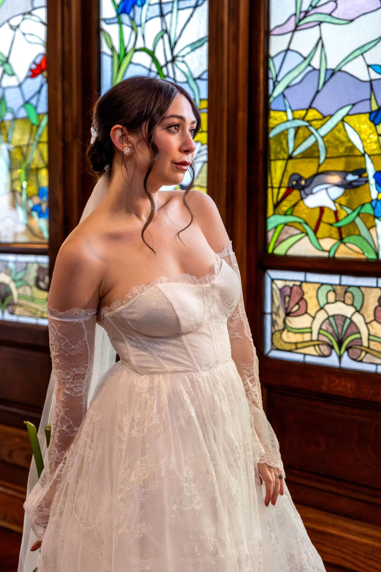 Bride in an off-shoulder lace wedding gown standing before colorful stained glass windows indoors.