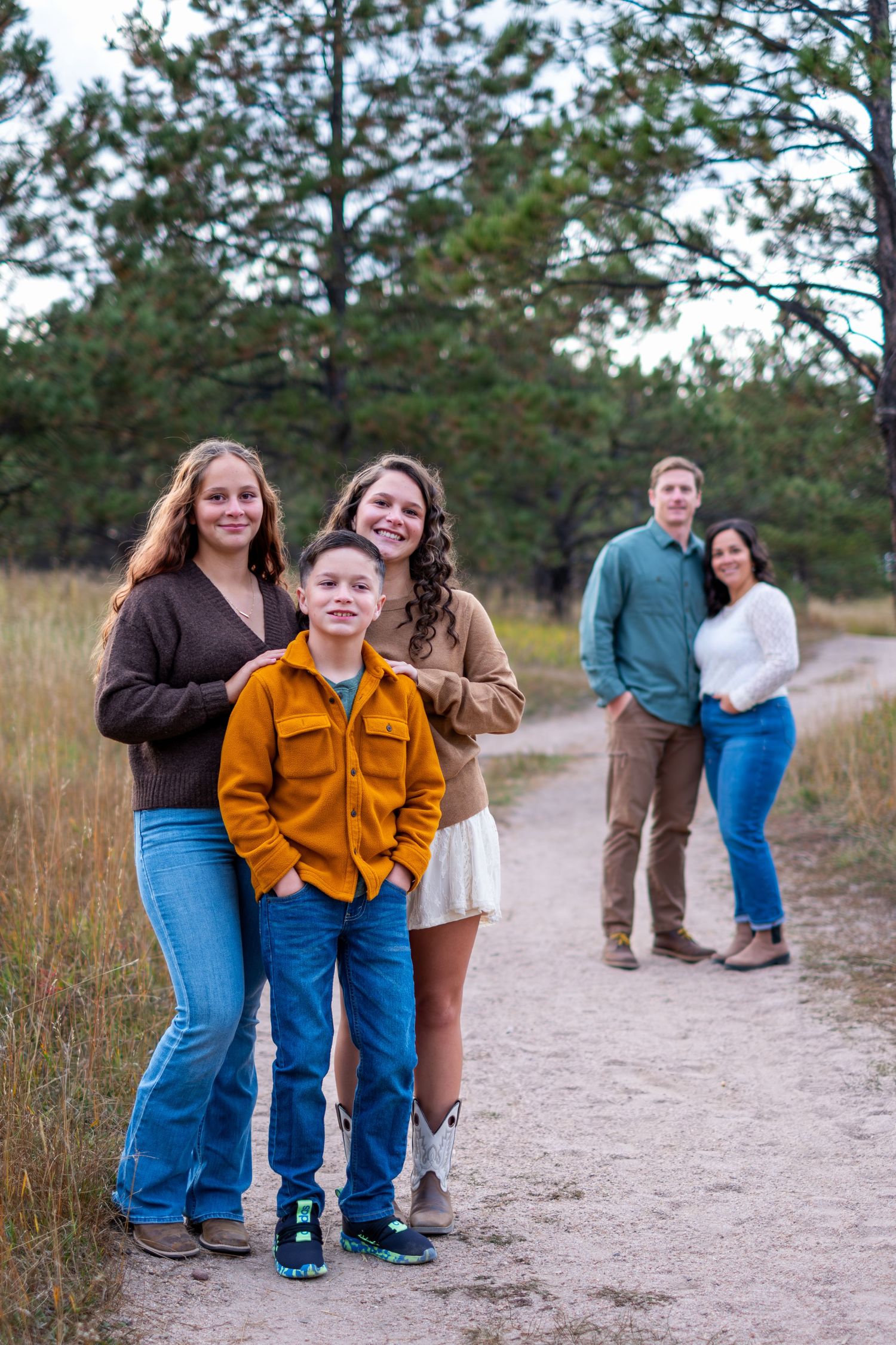 Family portrait with three siblings in foreground and parents on dirt path in pine forest.