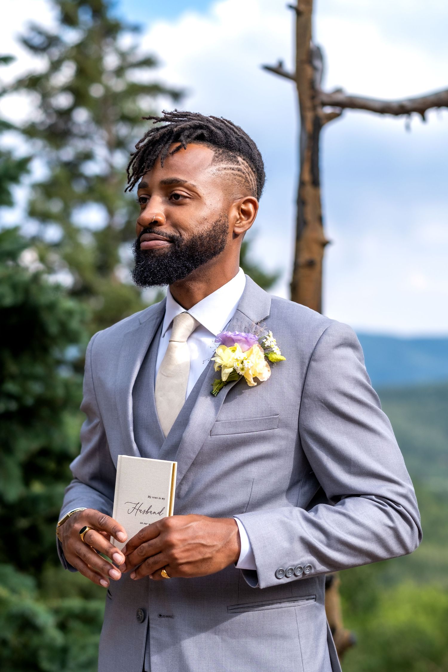 Handsome Black groom in gray suit holding vow booklet with boutonniere, standing outdoors with mountain forest backdrop.