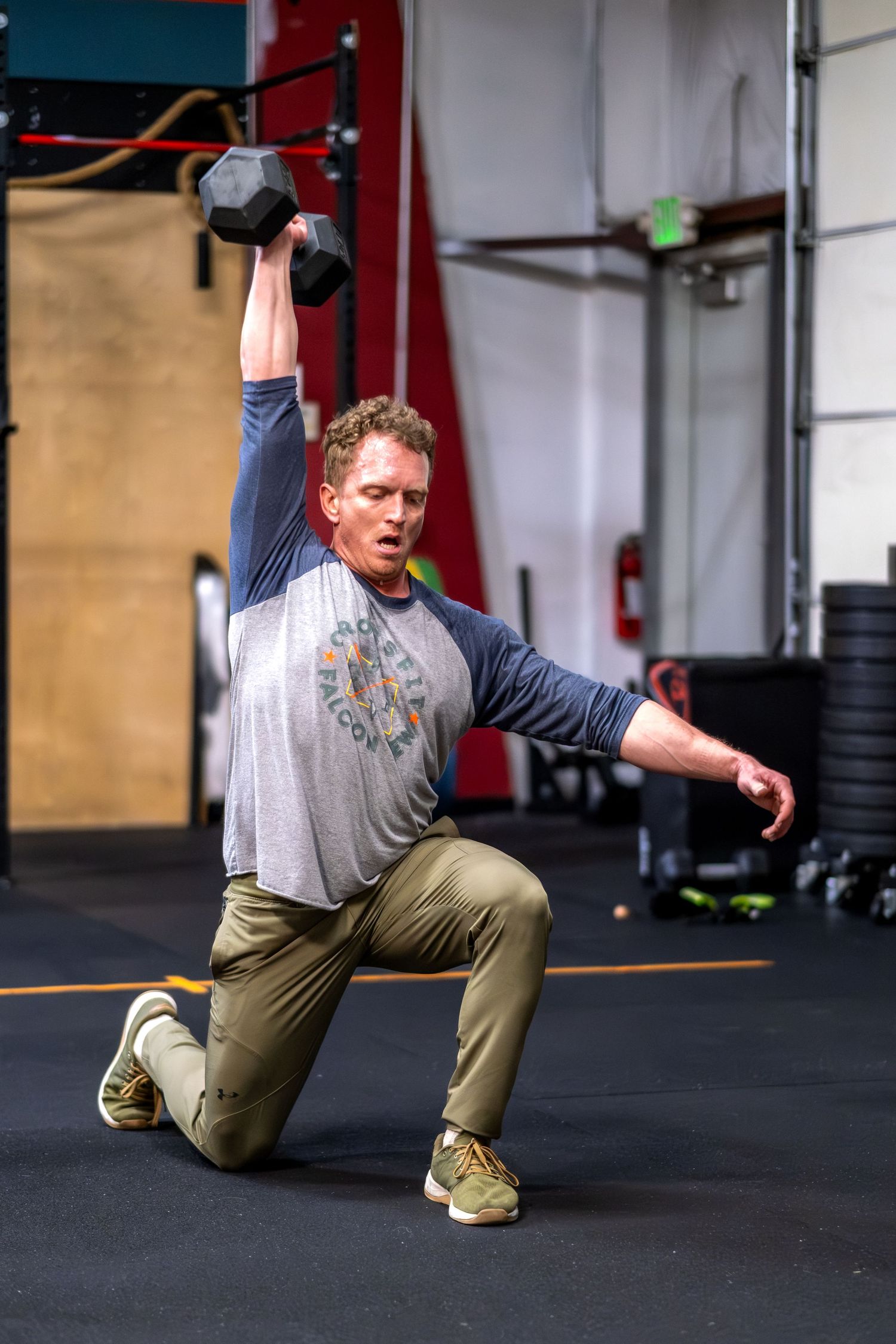 Redhead man in gray shirt performs a kneeling single-arm dumbbell overhead press in a gym.