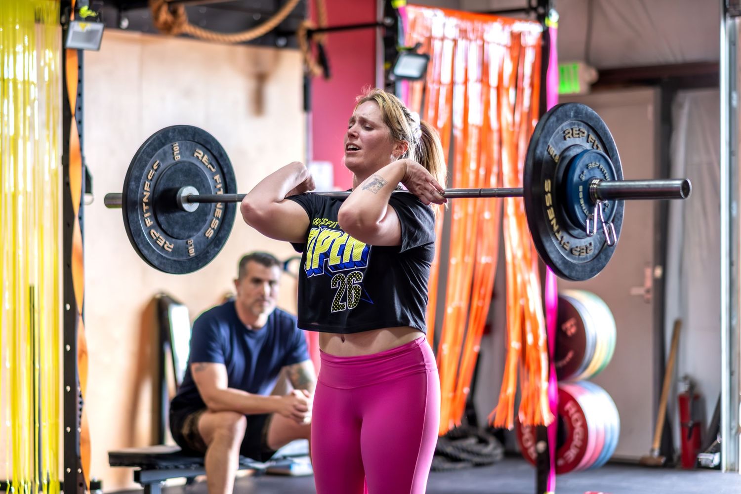 Blonde woman in pink leggings performing barbell clean lift at CrossFit gym.