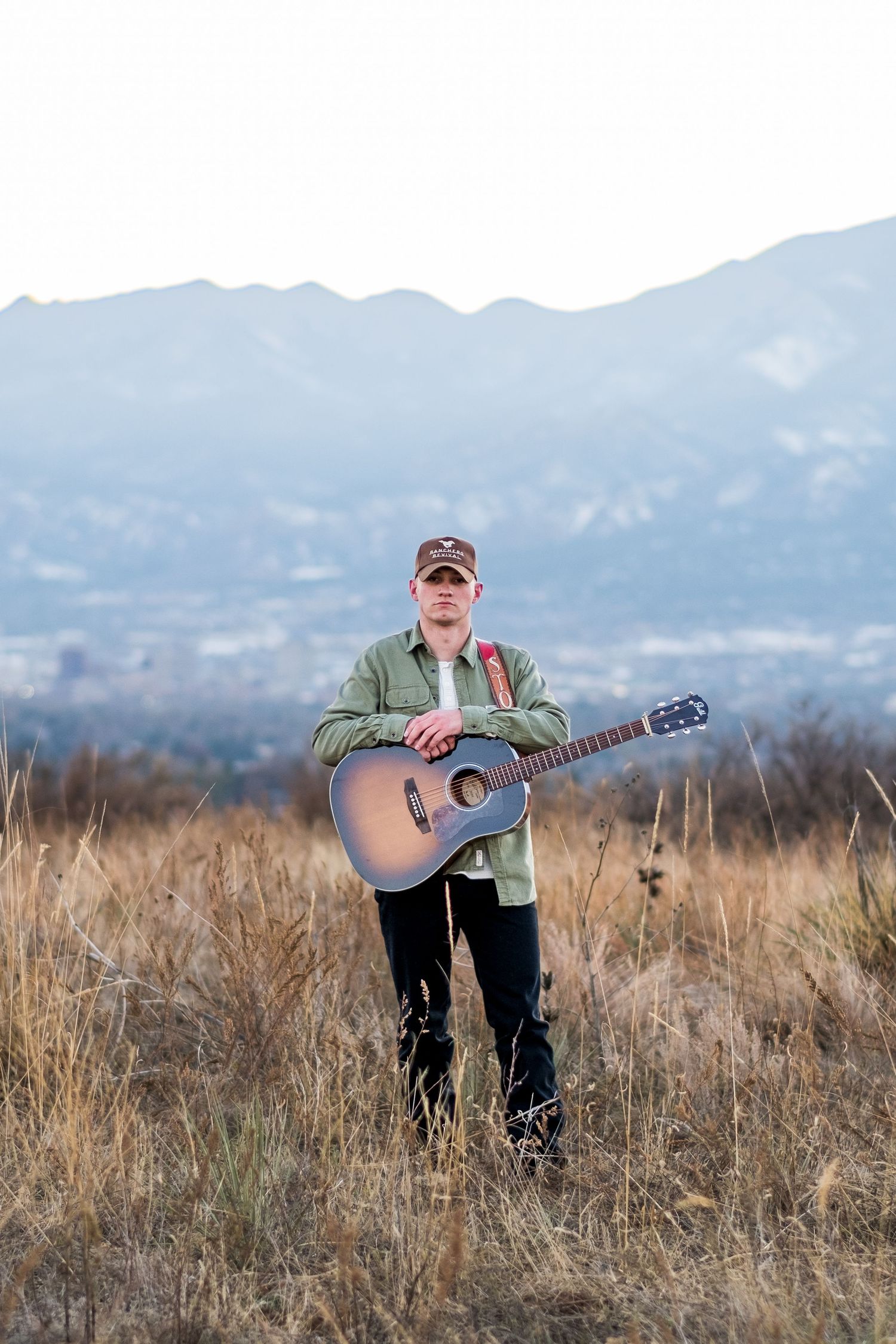 Young male guitarist standing in dry grass field with acoustic guitar and mountain backdrop at dusk.
