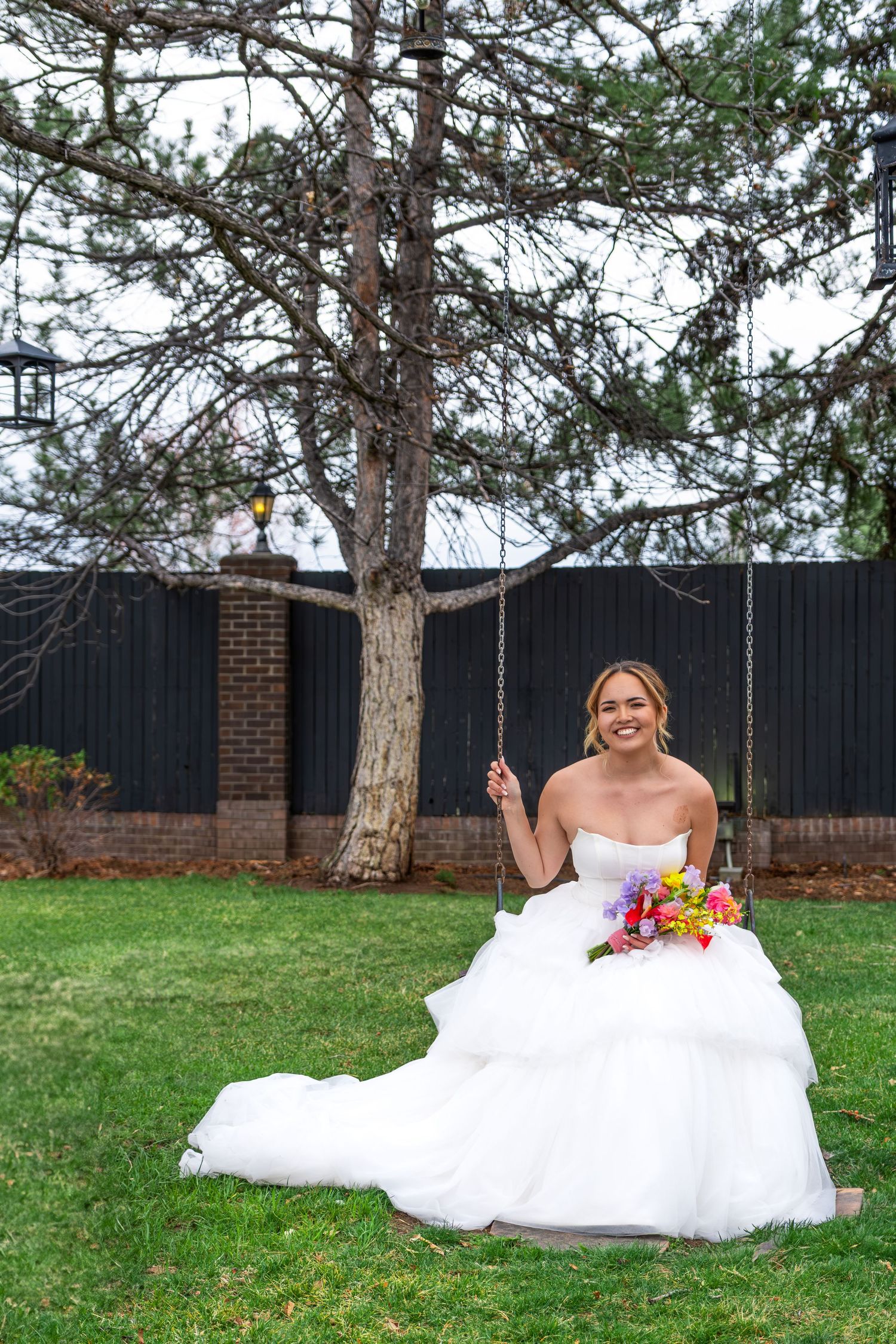 Smiling bride in strapless tiered gown sits on tree swing holding colorful bouquet in garden.