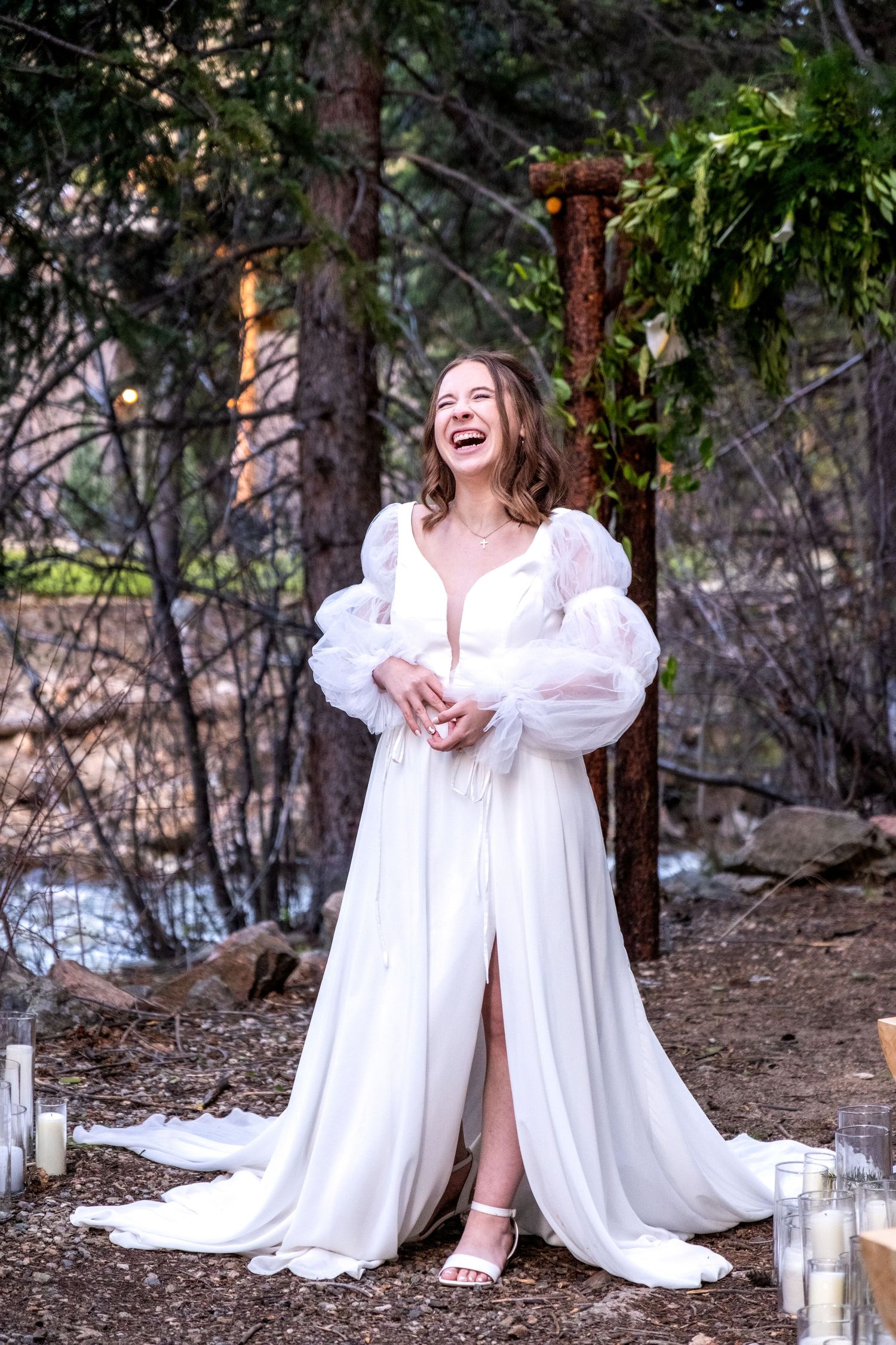 Laughing bride in white puff-sleeve wedding gown standing among candles beside rustic wooden arch in pine forest.