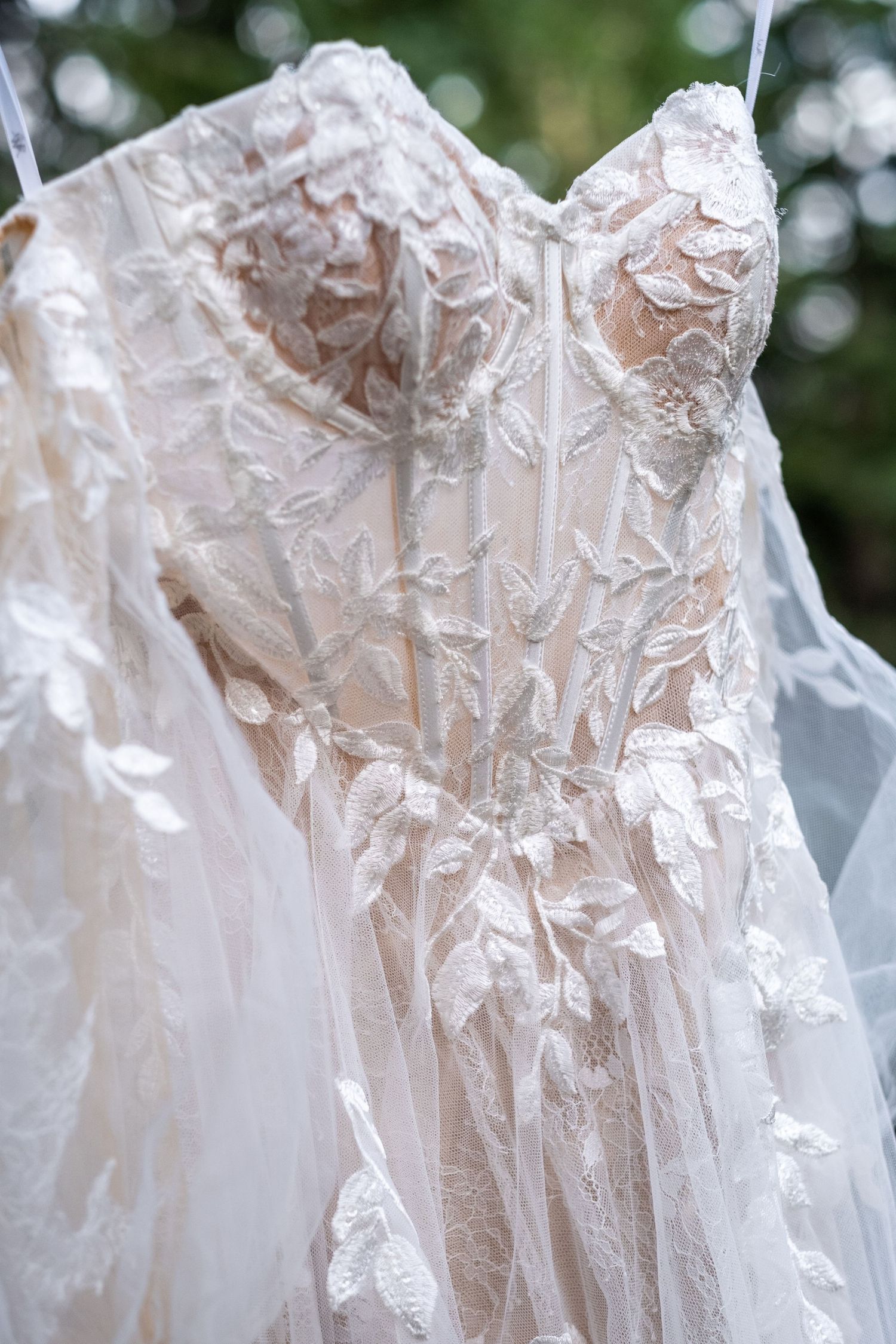 Elegant lace wedding gown with floral appliques hangs outdoors against a green bokeh background.