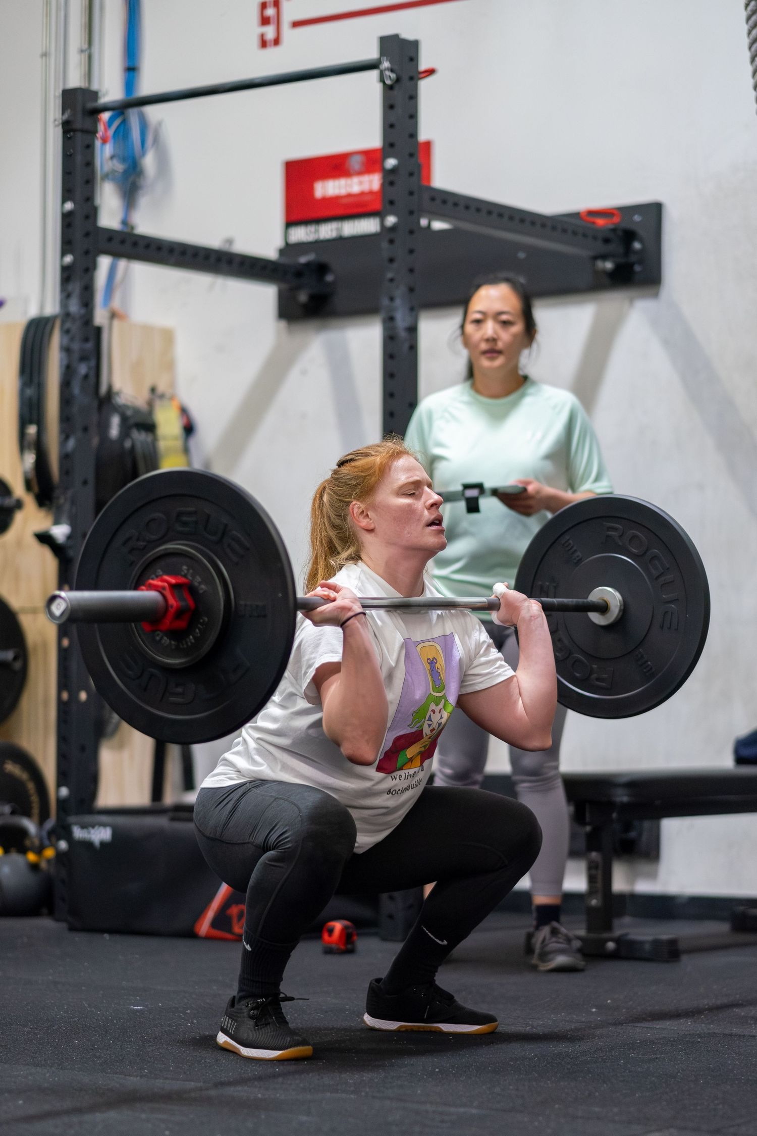 Young woman performs a barbell front squat while a coach monitors her form in a CrossFit gym.