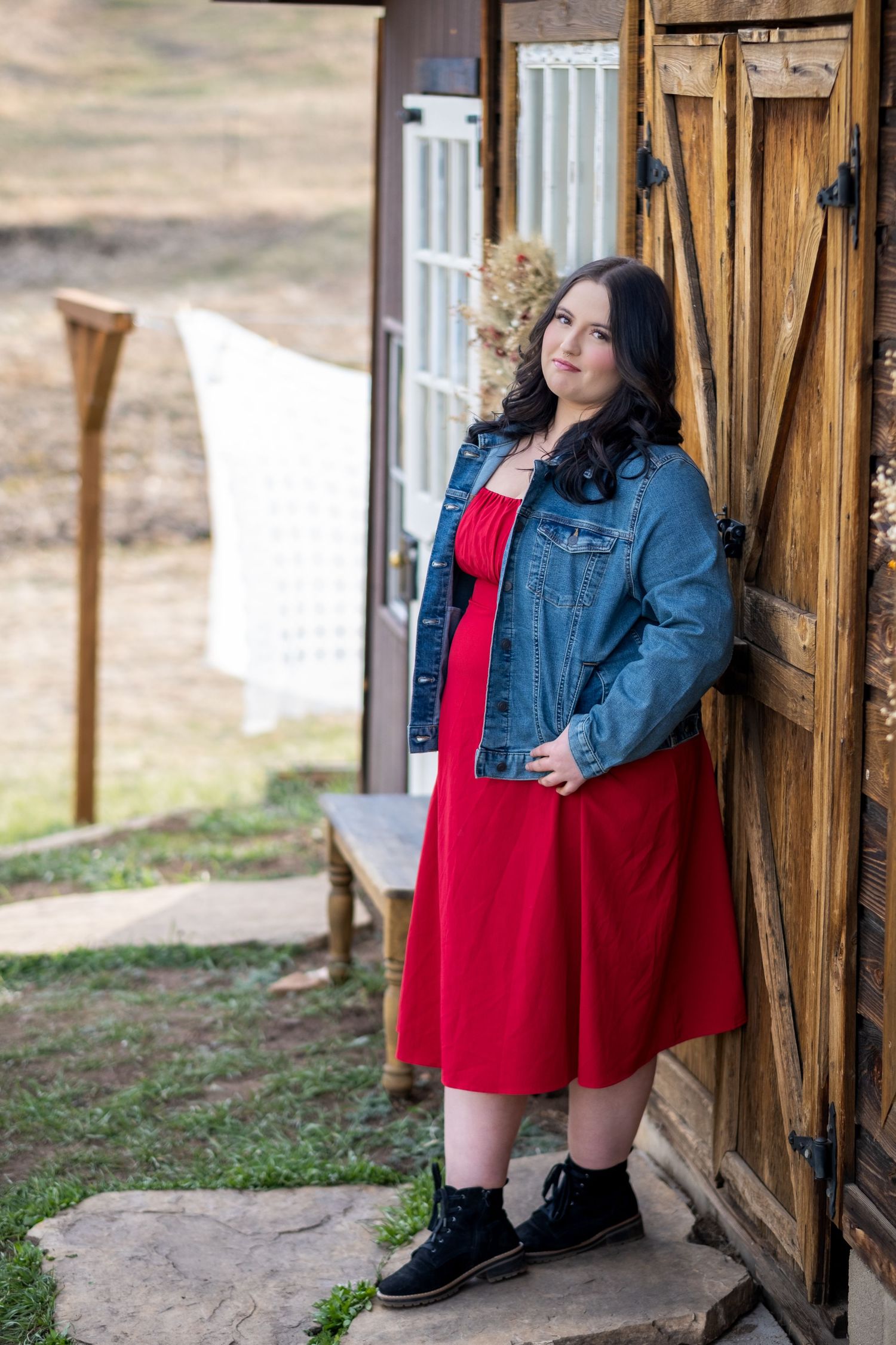Young woman in red dress and denim jacket posing beside a rustic wooden barn door outdoors.