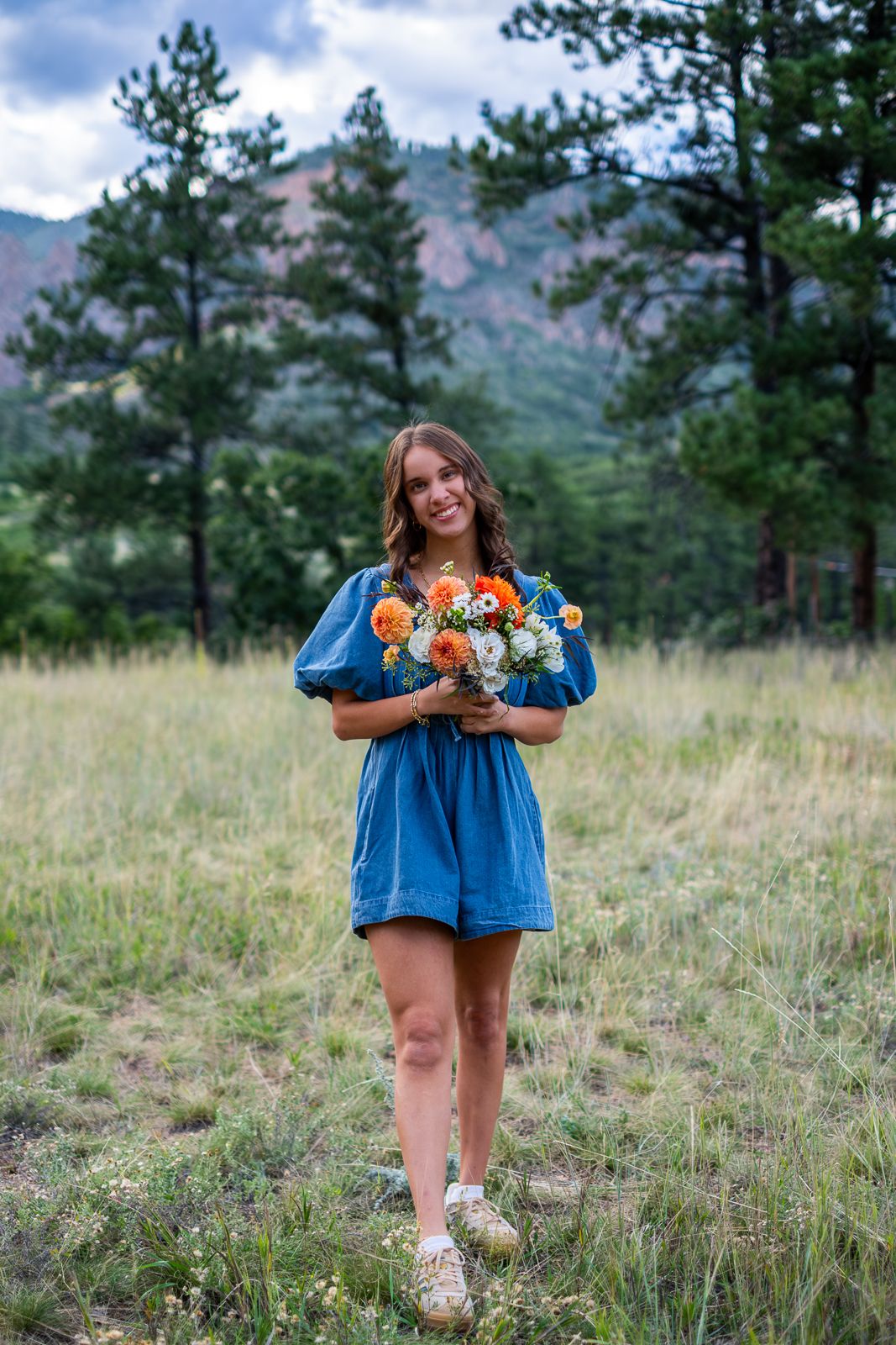 Smiling woman in blue dress holding colorful wildflower bouquet in mountain meadow.
