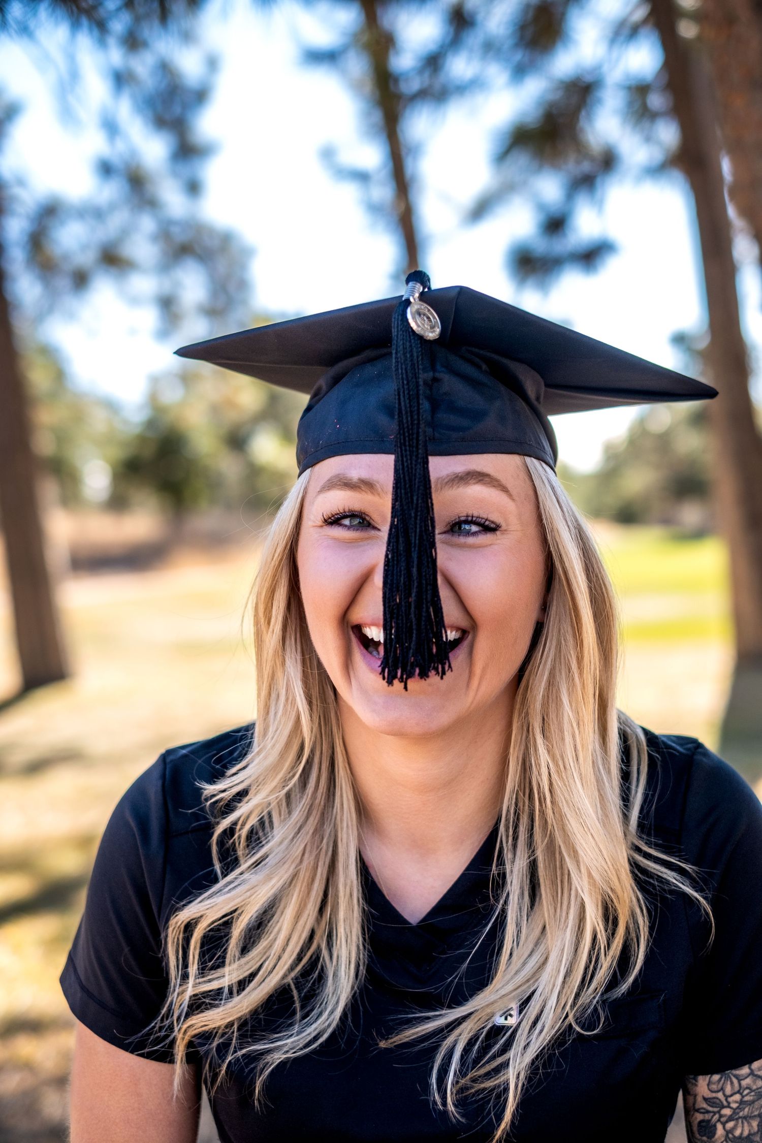 Laughing blonde woman wearing black graduation cap outdoors among tall trees.