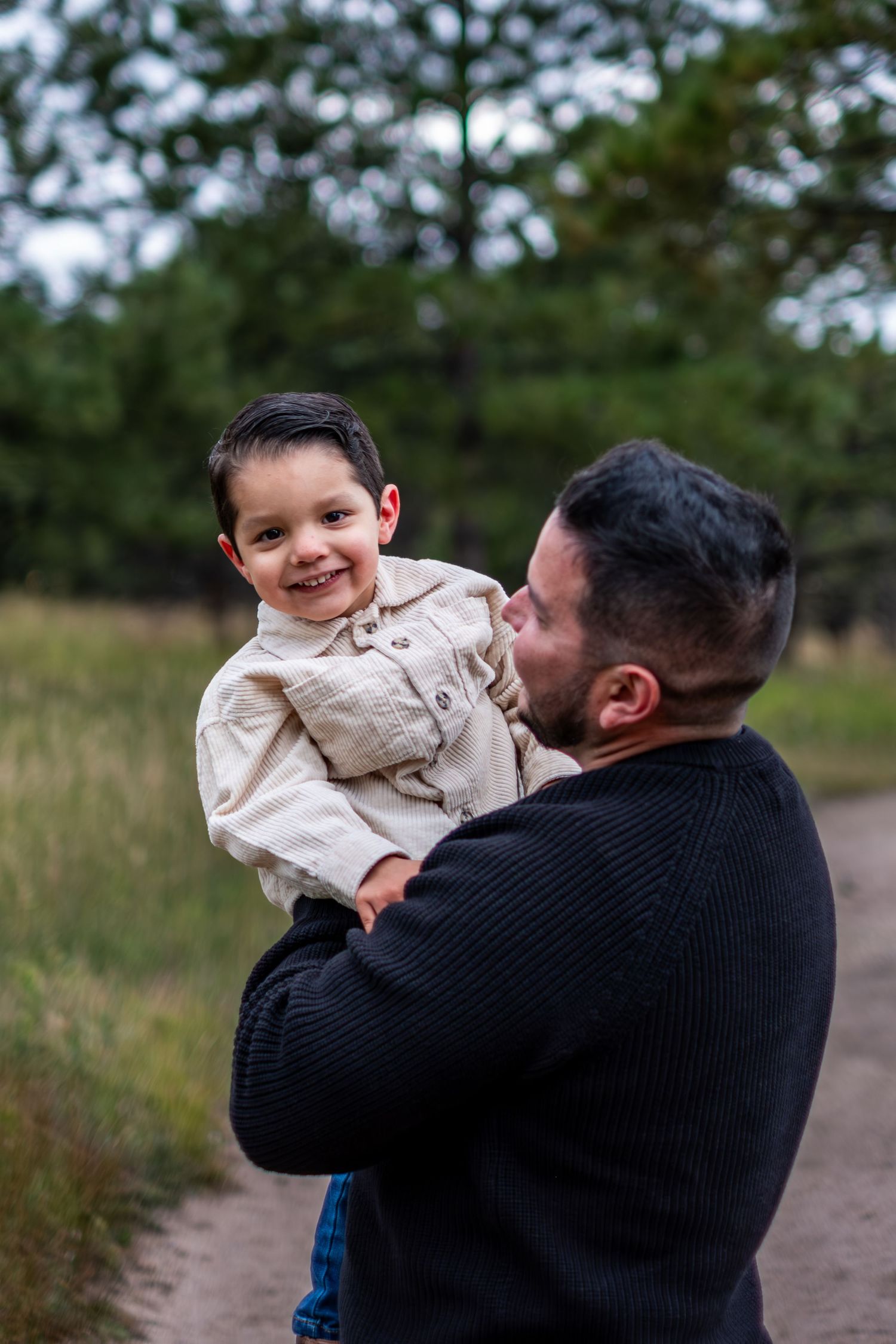 Smiling toddler being held by father outdoors on a forest path surrounded by trees.