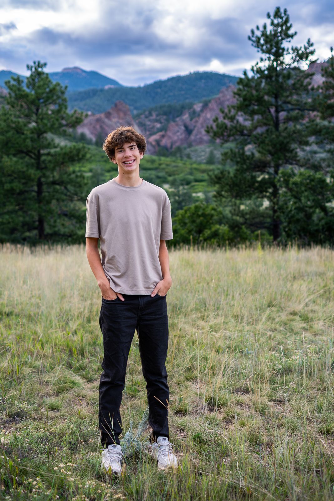 Smiling teenage boy standing in a Colorado mountain meadow with pine trees and red rock formations in background.