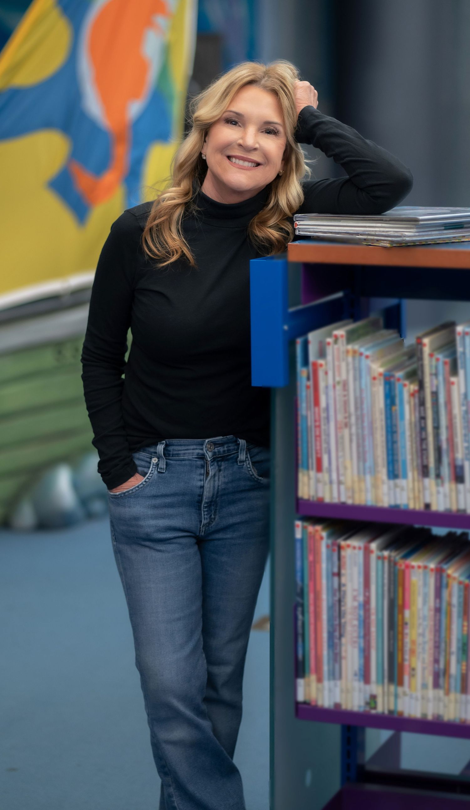 Smiling female author with her arm propped up on a library shelf surrounded by children's books