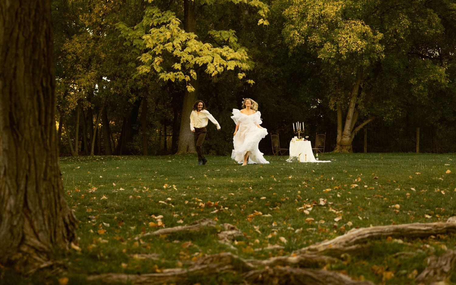 Bride and groom running playfully through a lush green park scattered with autumn leaves.