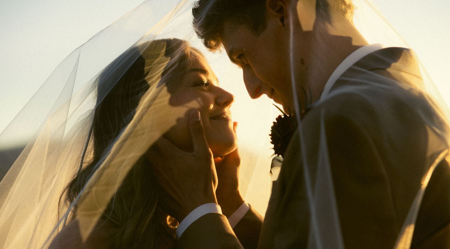 Romantic backlit portrait of bride and groom under a veil at golden hour sunset.