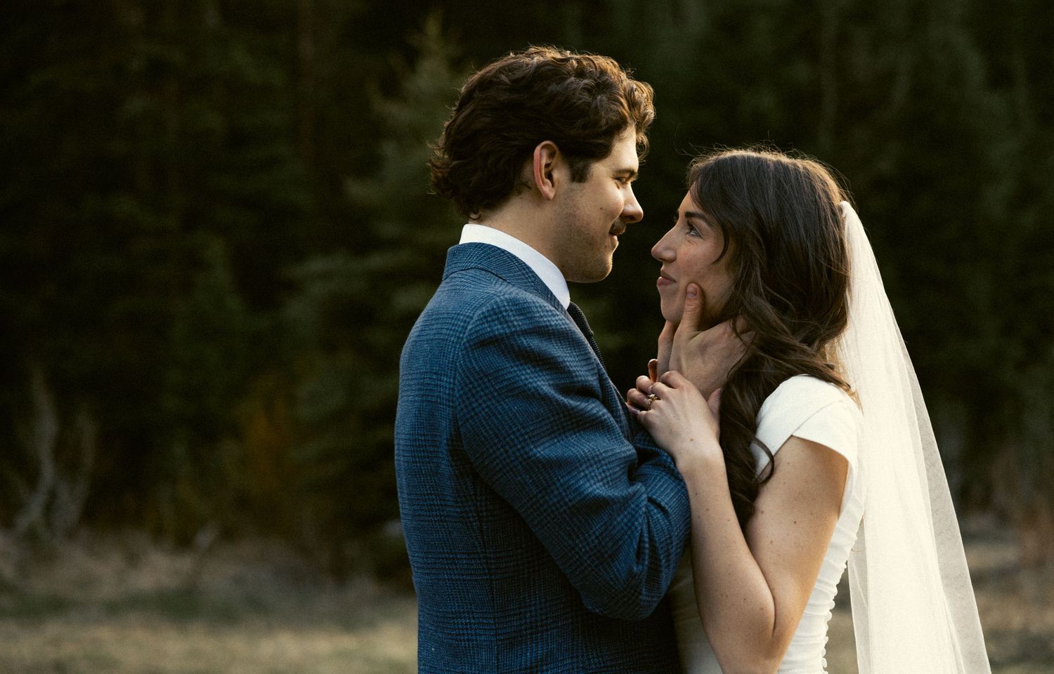 Bride and groom share an intimate moment in a forest clearing, groom in blue suit with veil.