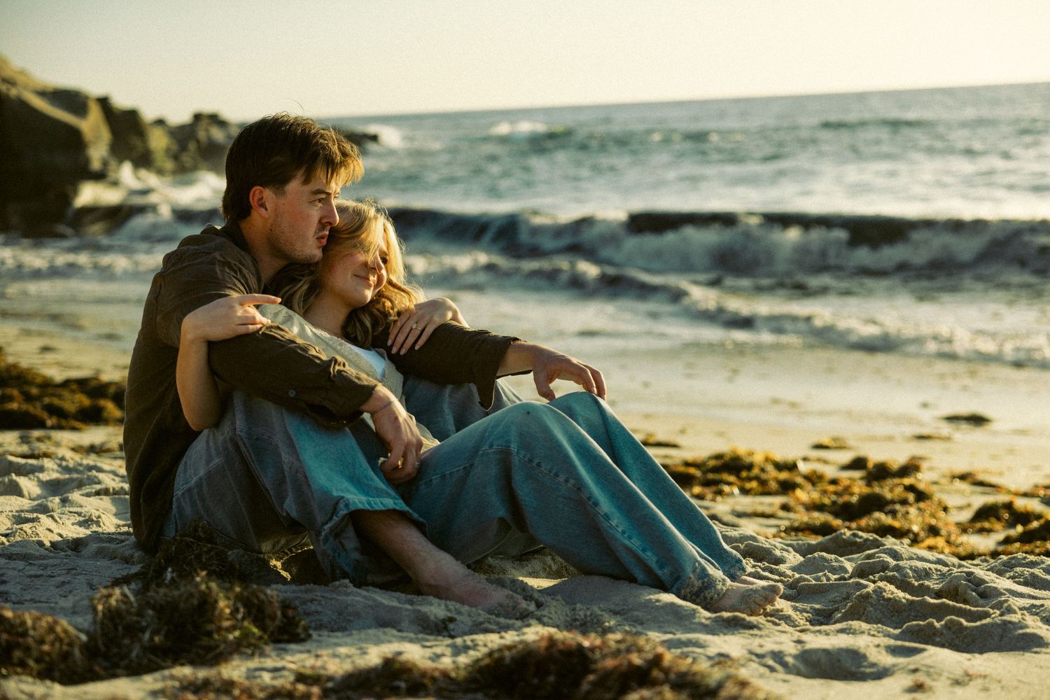 Couple sitting together on a sandy beach at sunset, embracing romantically with ocean waves in the background.