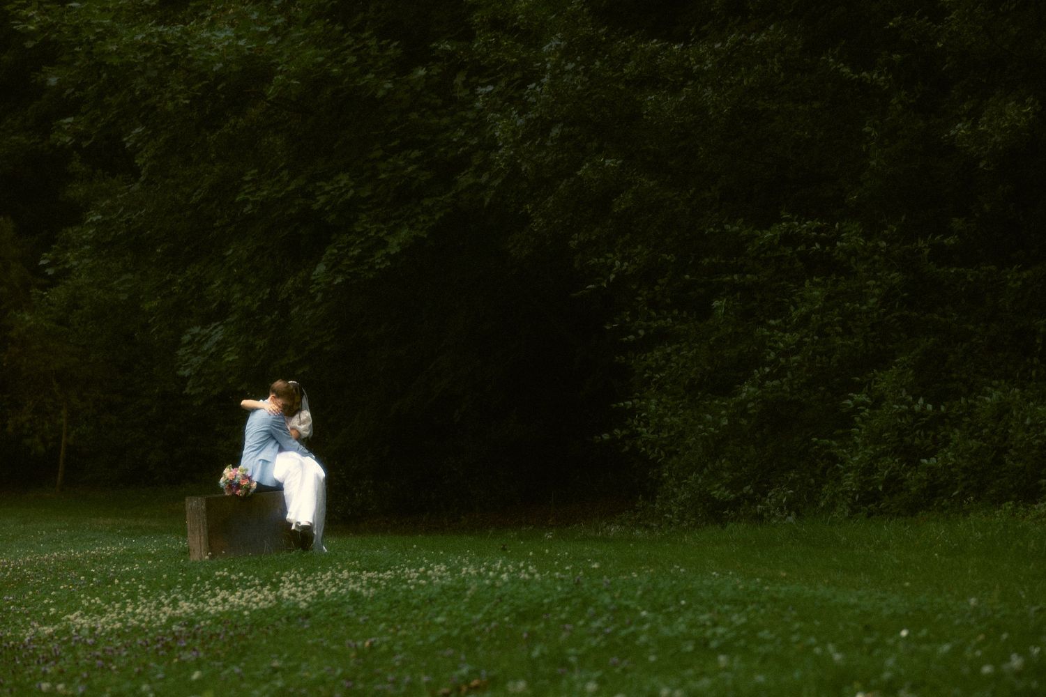 Bride sitting alone on a suitcase in a sunlit meadow surrounded by a lush dark tree line in Switzerland.