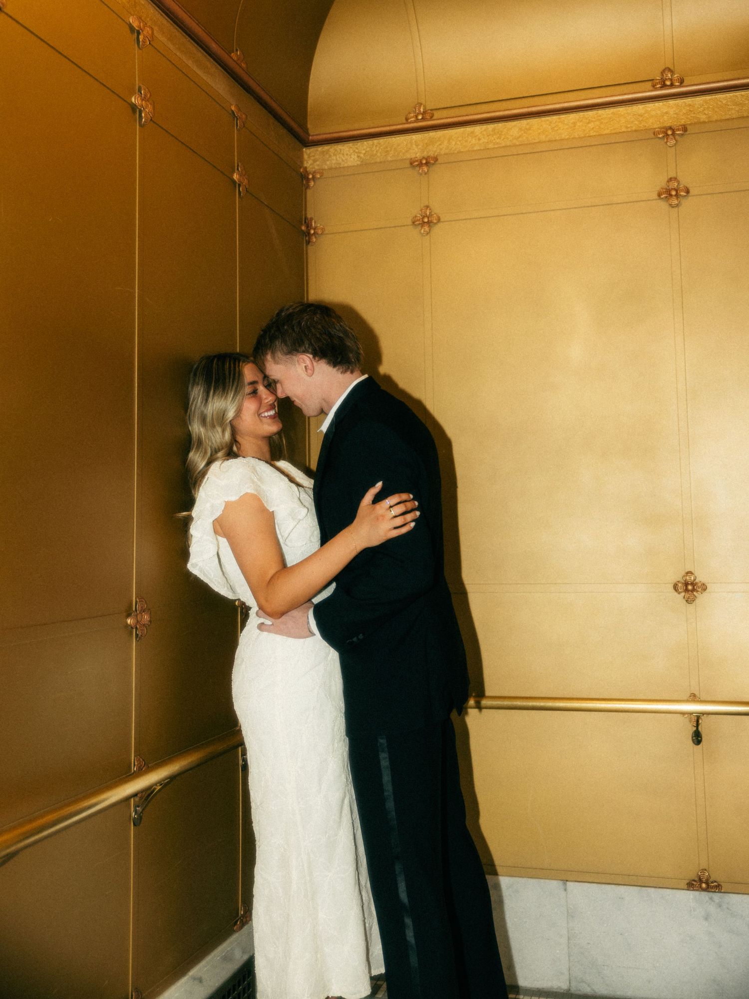 Bride in white dress and groom in black suit share a romantic moment against a gold wall.