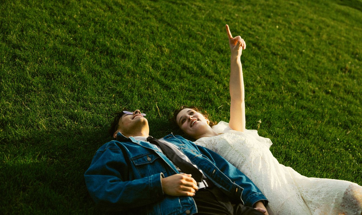 Couple lying on green grass, woman in white dress pointing up at the sky.