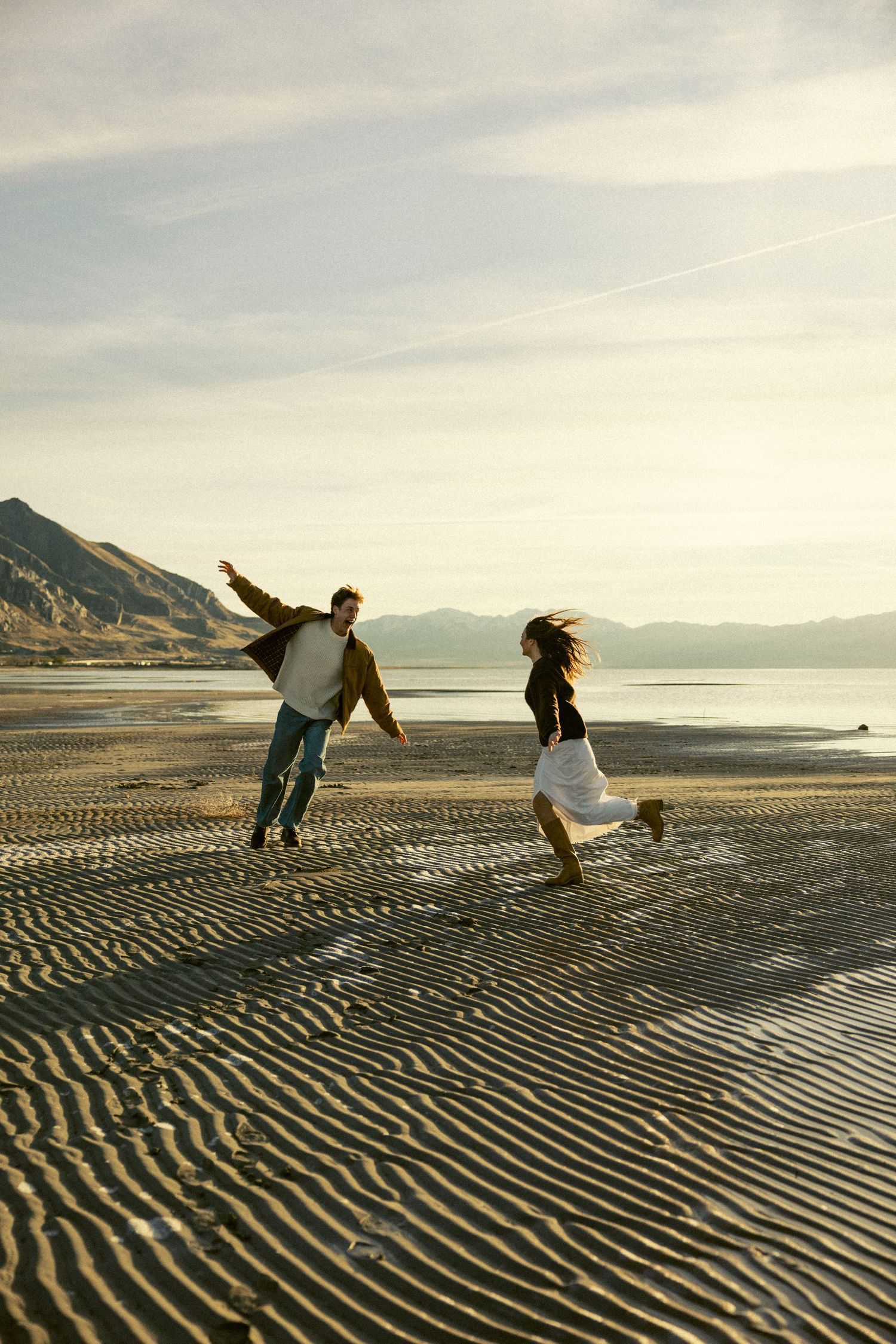 Couple playfully running on a sandy beach at sunset with mountains in the distance.