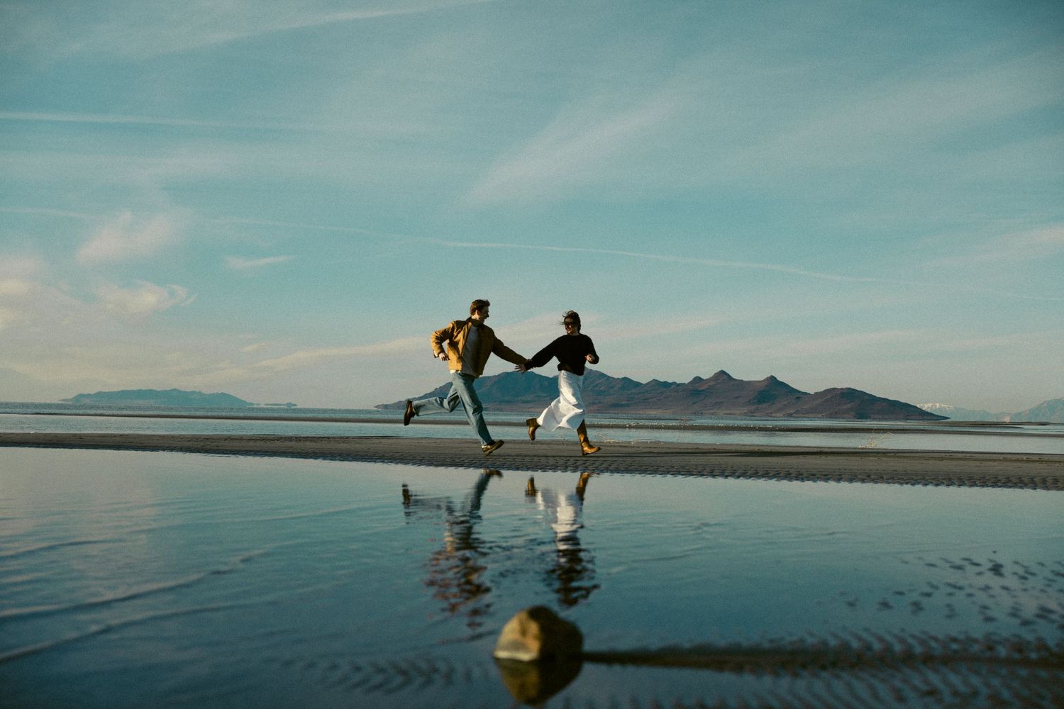 Couple holding hands running joyfully on a reflective beach with mountains in the background.
