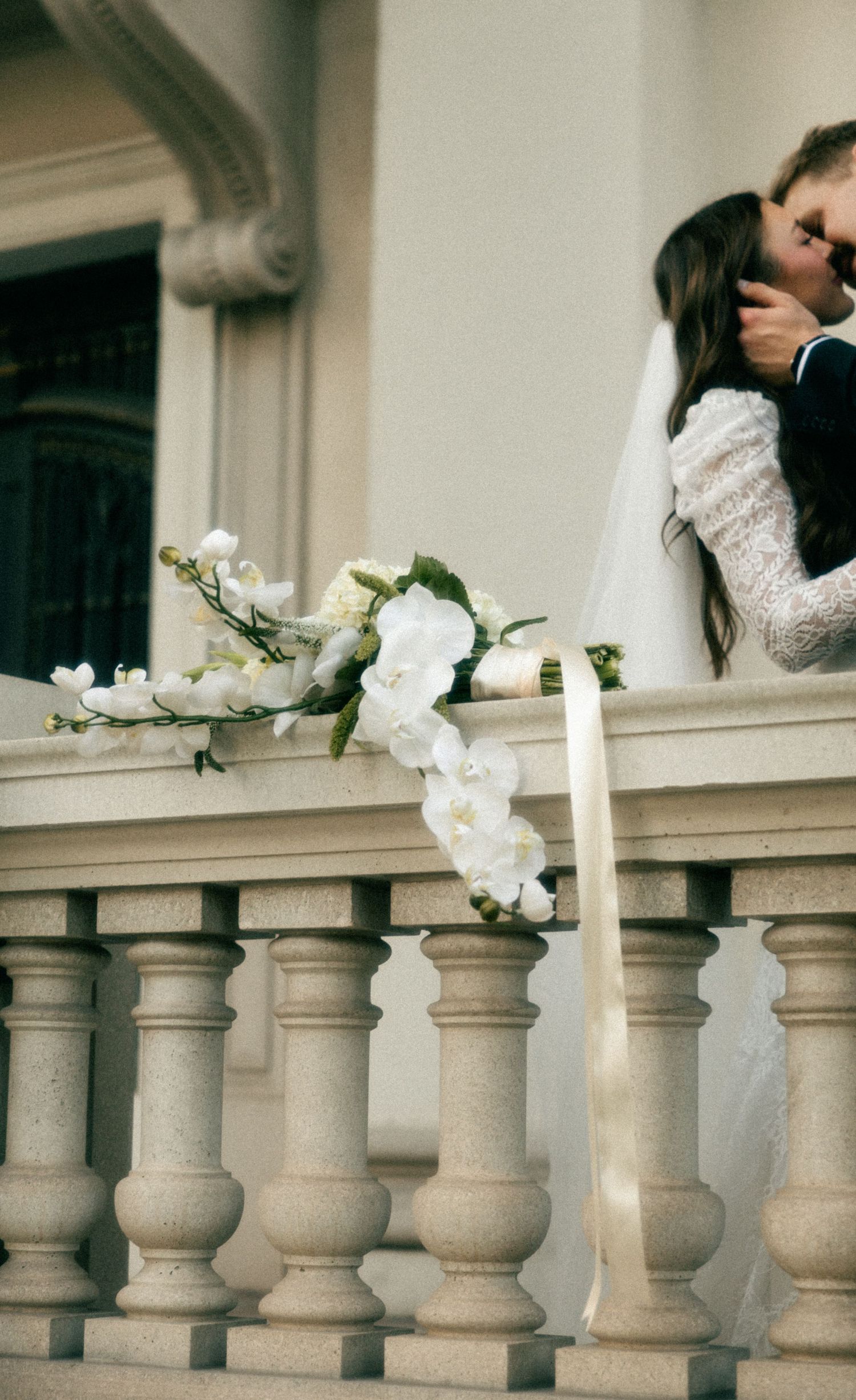 Bride and groom sharing a kiss on a stone balustrade with a cascading white orchid bouquet.