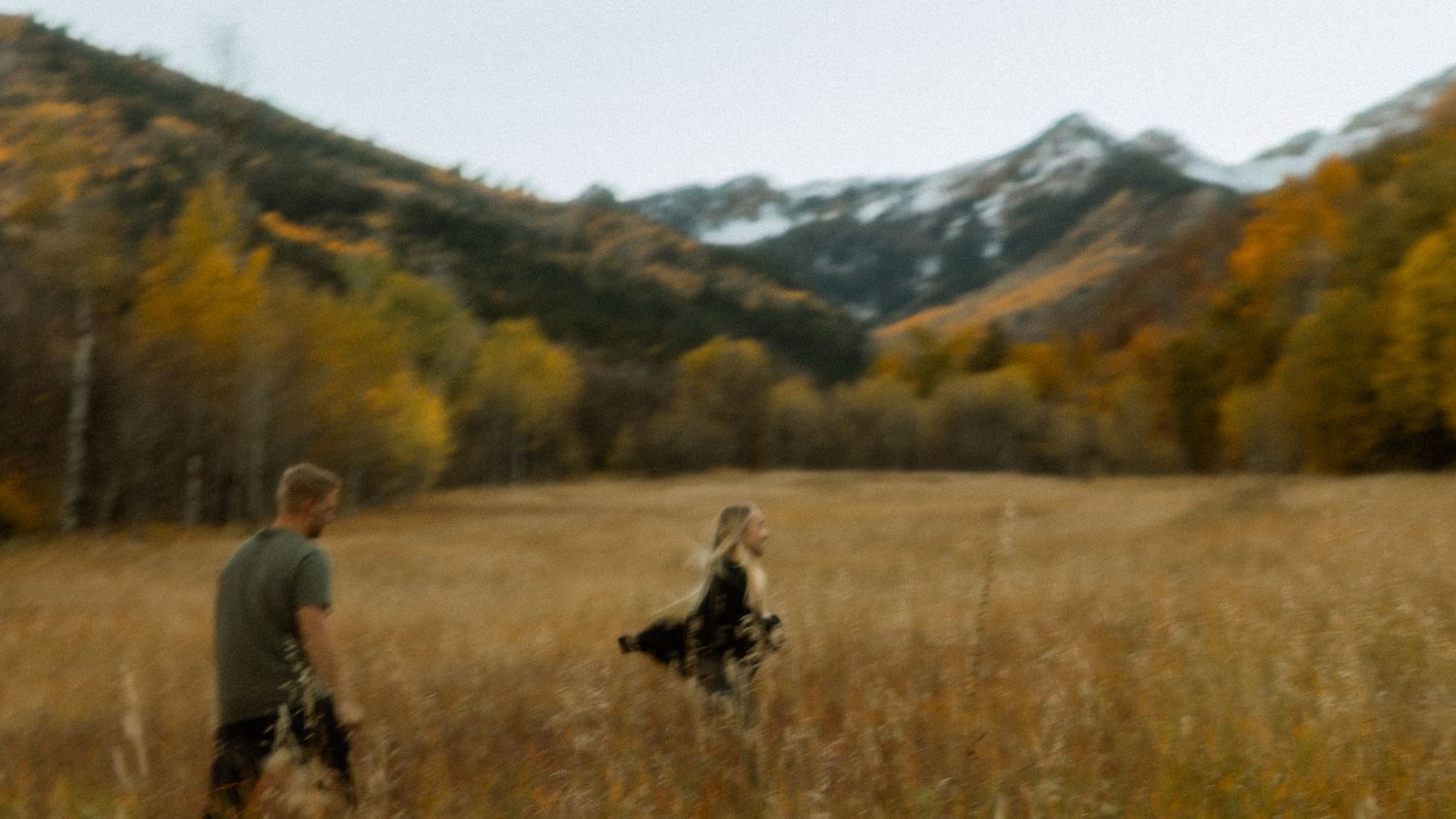 Couple walking through golden autumn meadow with snow-capped mountains in background.