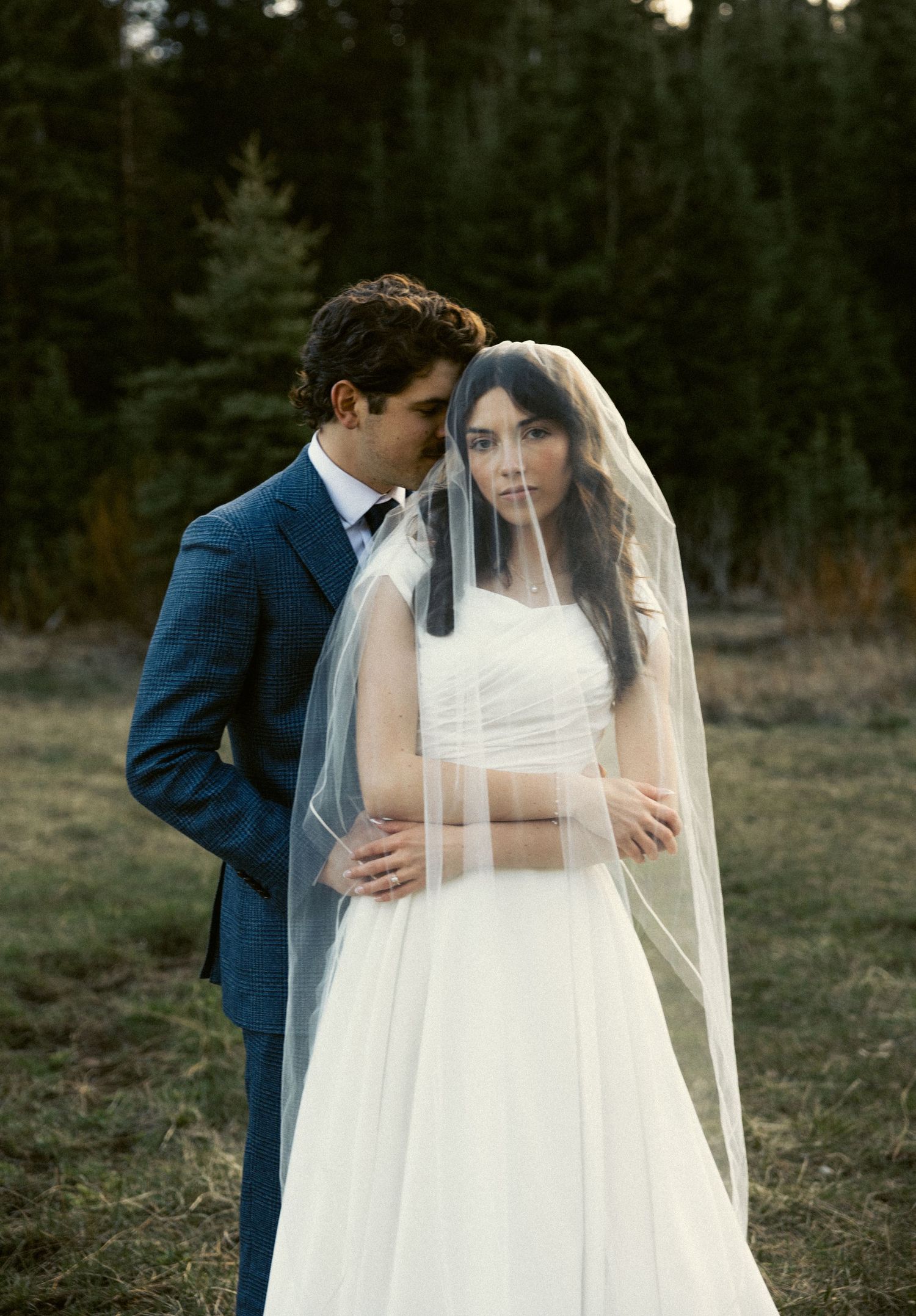 Groom in blue suit embracing bride in white gown and veil in a forest meadow at dusk.