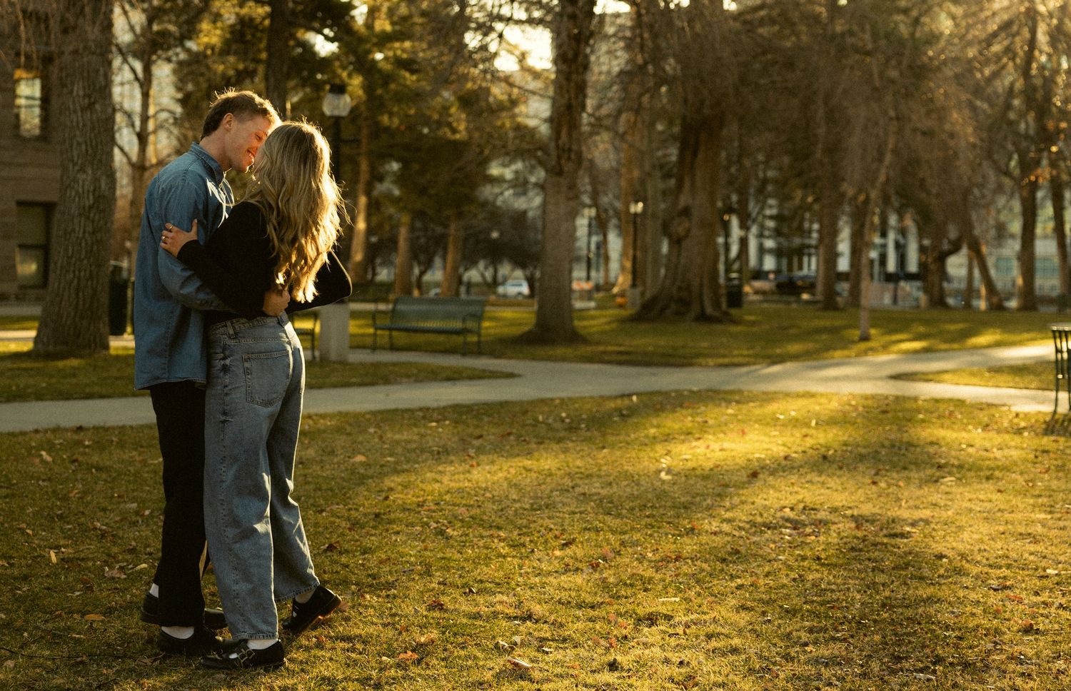 Young couple sharing a tender kiss in a sunlit park surrounded by tall trees.