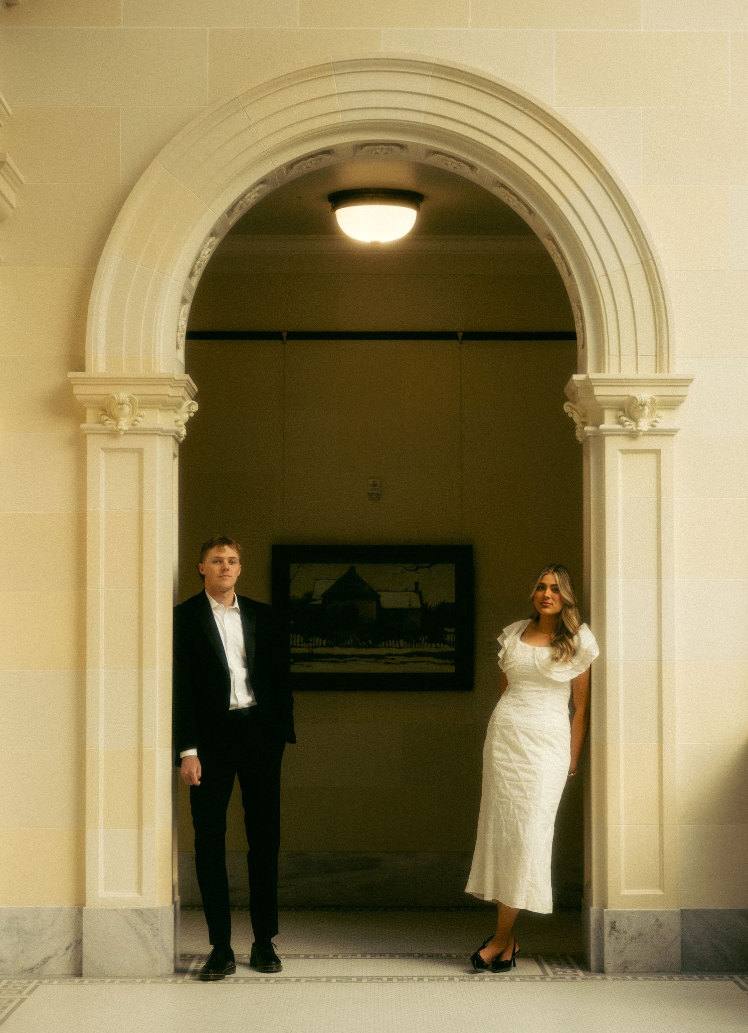 Couple in wedding attire standing beneath an elegant stone archway in a classical building interior.