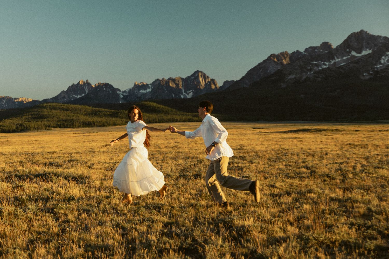 Couple in white wedding attire running through golden meadow with snow-capped mountains at sunset.