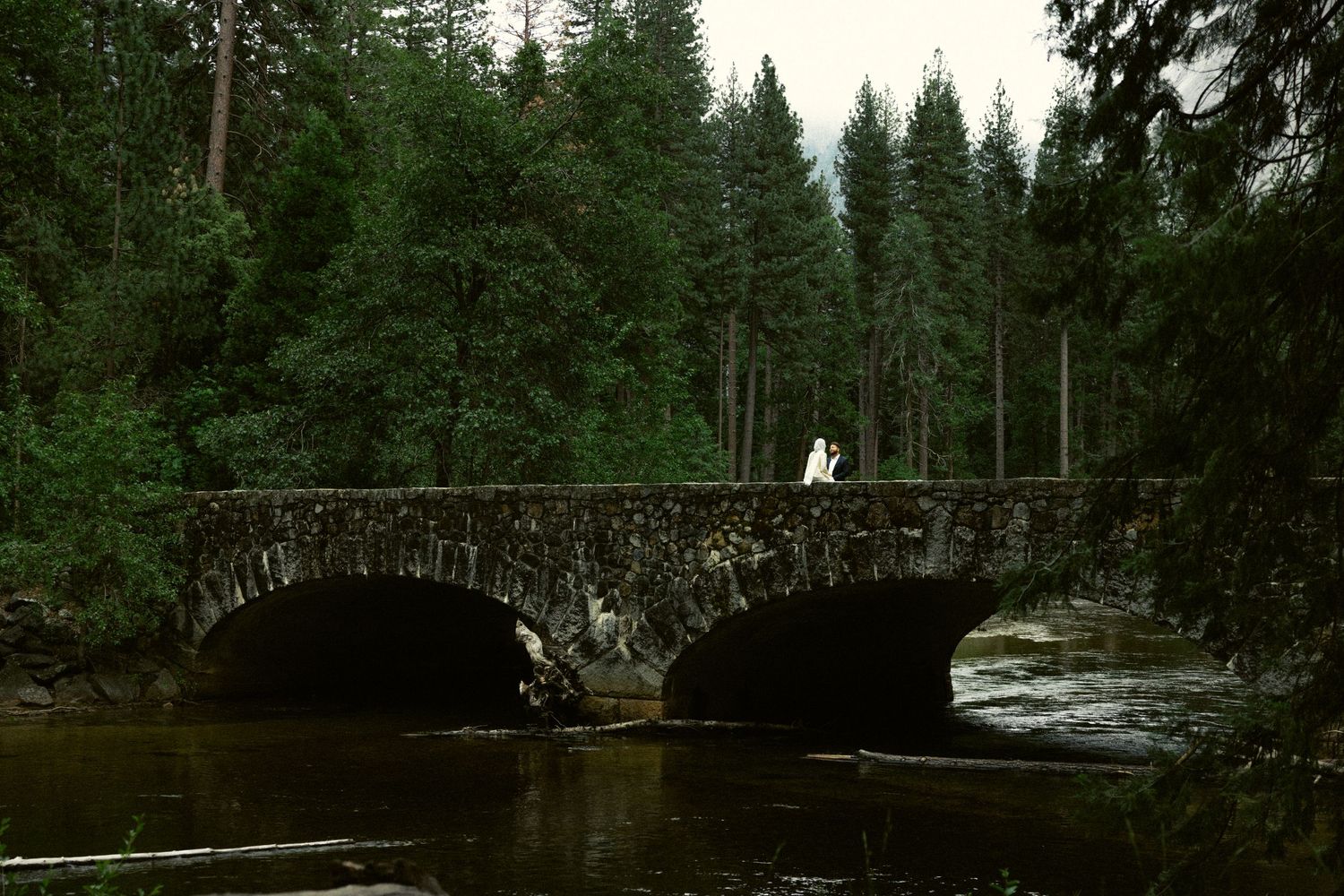 Bride and groom standing on a rustic stone arched bridge over a river in a dense forest in Yosemite National Park.