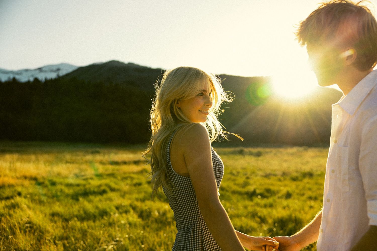 Blonde woman smiling over shoulder holding hands with partner in a sunlit mountain meadow.