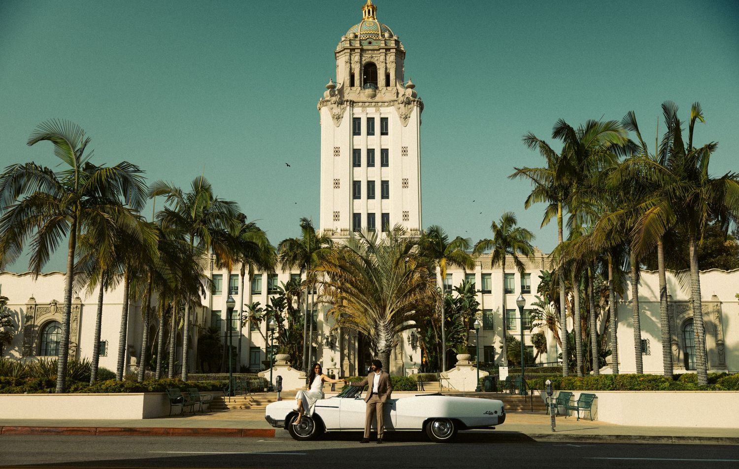 Couple posing near vintage cars in front of Beverly Hills City Hall lined with tall palms.