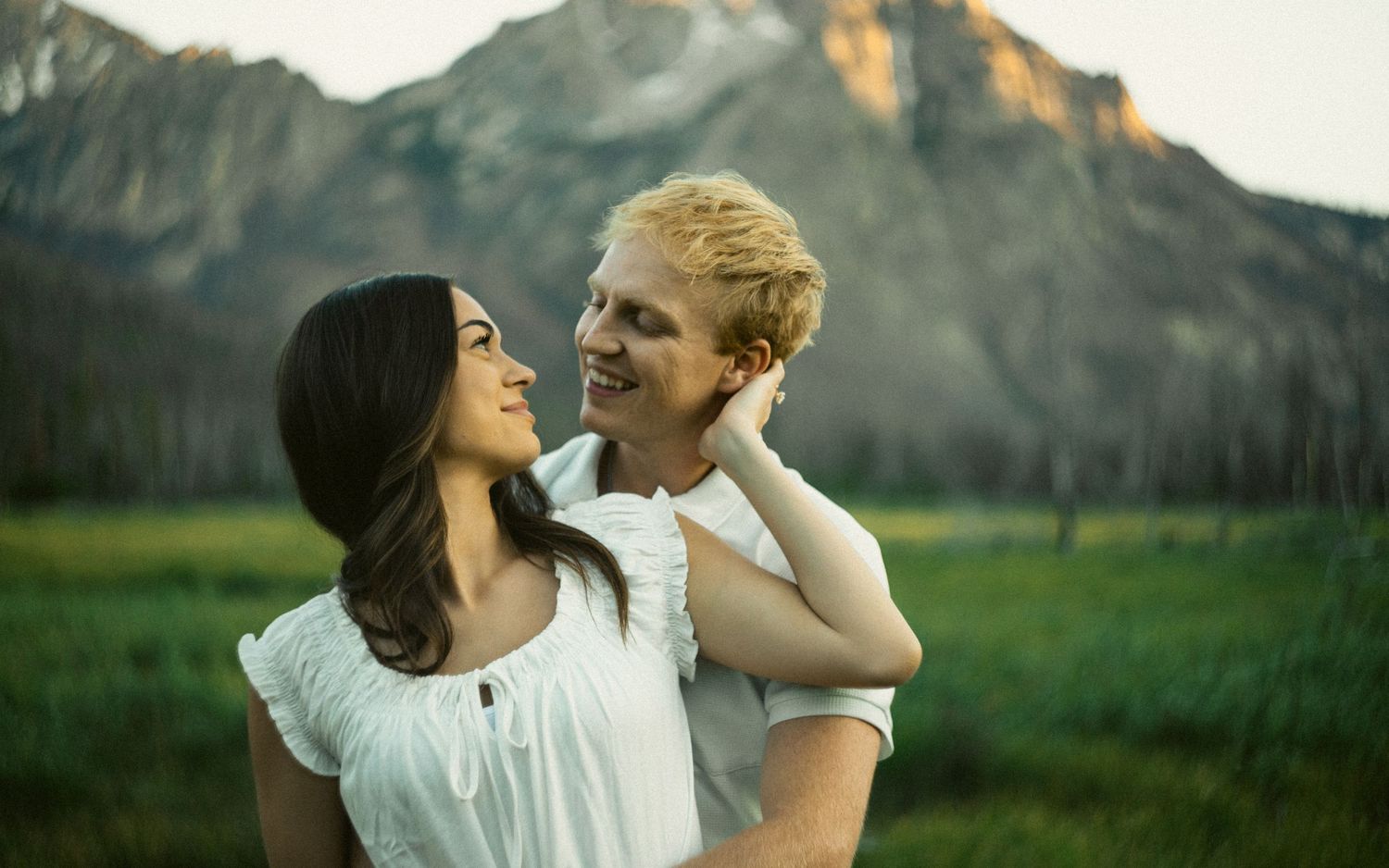Smiling couple in a green meadow with a dramatic mountain peak glowing in background.