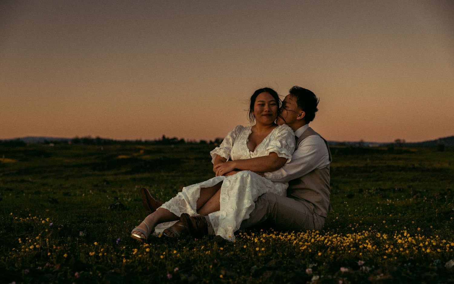 Couple sitting together in a California flower field at sunset with a warm glowing sky.
